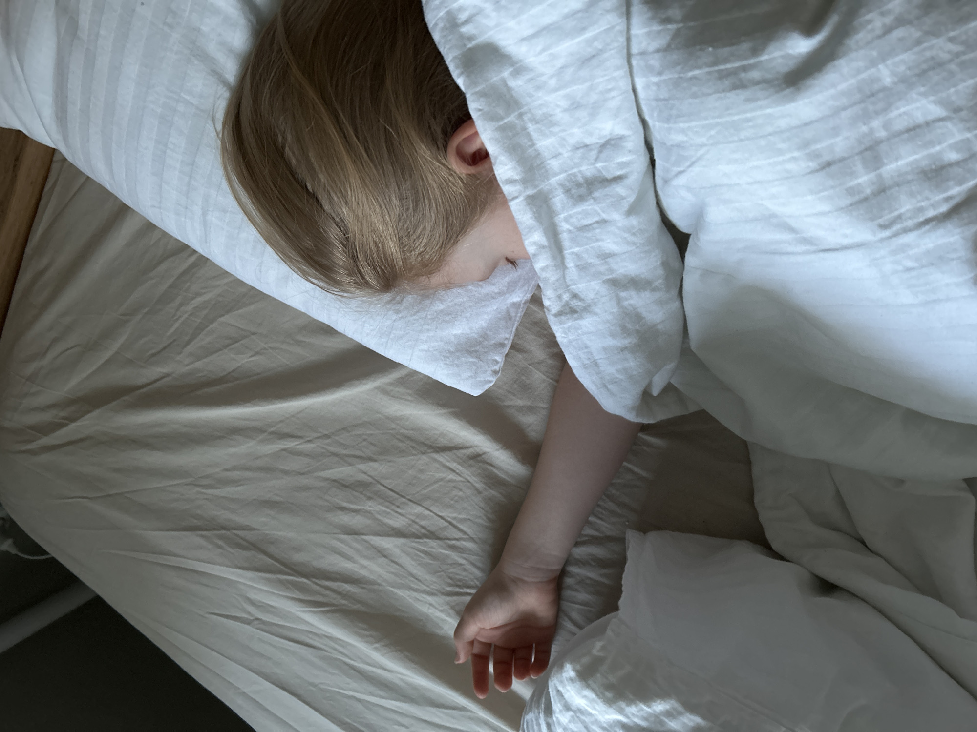 The image presents a top-down view of a person sleeping peacefully in a bed. The subject, likely a child or young adult with light brown or blonde hair, has their head resting on a white pillow with subtle stripes. Most of their face is obscured by their hair and the pillow, with only an ear and a small portion of their cheek visible. Their body is largely covered by a white duvet or comforter, appearing soft and rumpled. One arm extends from beneath the covers, resting on a light beige fitted sheet, with the hand gently curled. A part of a wooden bed frame is visible on the left side. The soft, natural lighting suggests it is daytime, possibly morning. The overall atmosphere conveys tranquility and rest. No specific location cues are visible within the image, but the location context is Chaikovsky, Russia. There is no visible text.