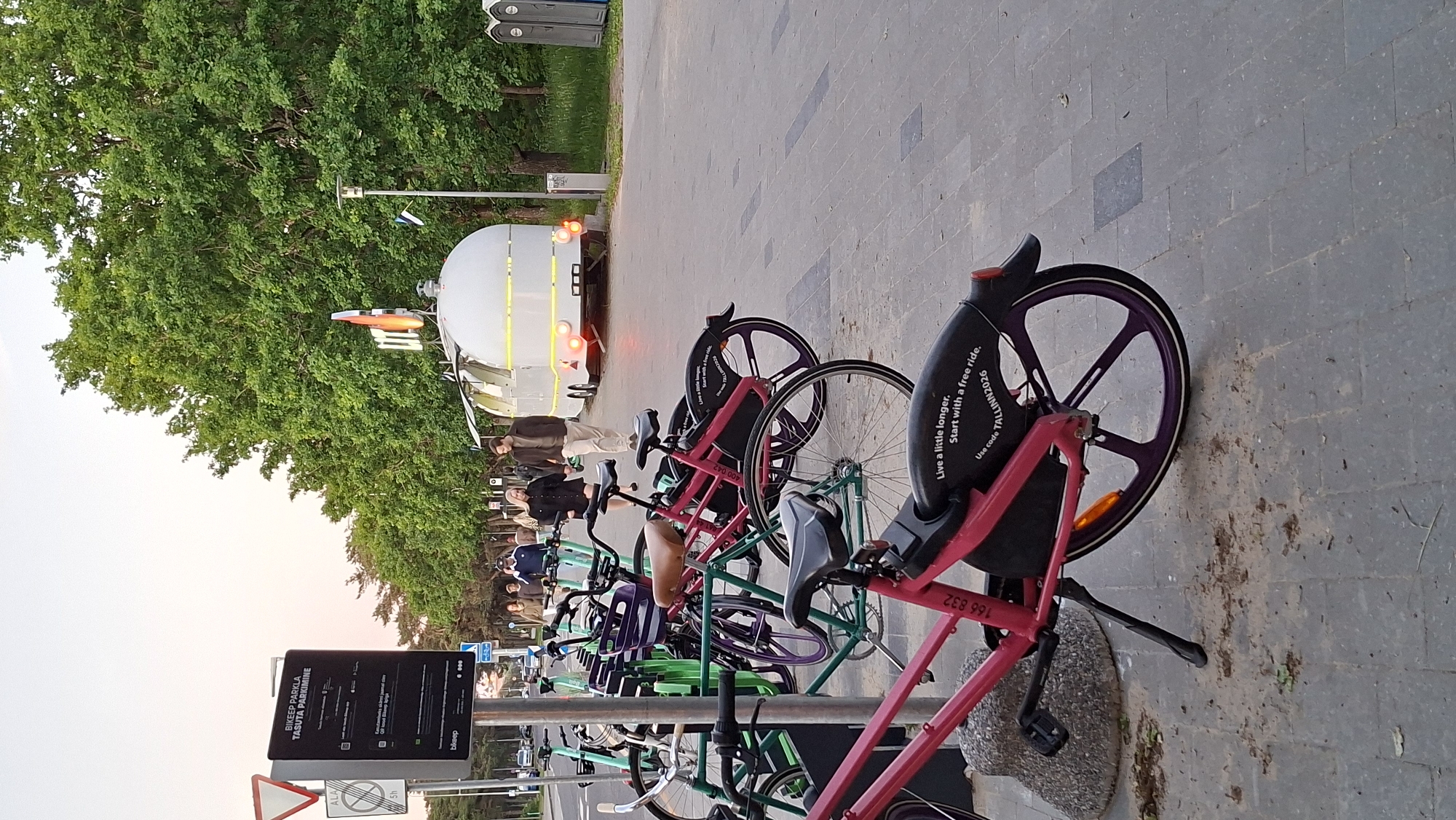The image depicts a street scene in Tallinn, Estonia, possibly in the Kadriorg district, known for its parks and coastal areas. In the foreground, a row of parked bicycles, predominantly pink and green, is visible. Some of the bicycles have informational stickers on their rear fenders, one of which reads "Live a little longer. Start with a free ride. Use code TALLINN2026." Behind the bicycles, a white food truck with the word "Ülo" on its signage is parked. Several people are walking along a paved walkway beside the parked bicycles and the food truck. The walkway is lined with trees, and in the distance, there are portable toilets. The sky is overcast, suggesting it might be late afternoon or early evening. The overall impression is a public space in Tallinn where residents and visitors can access bicycle rentals and food services.