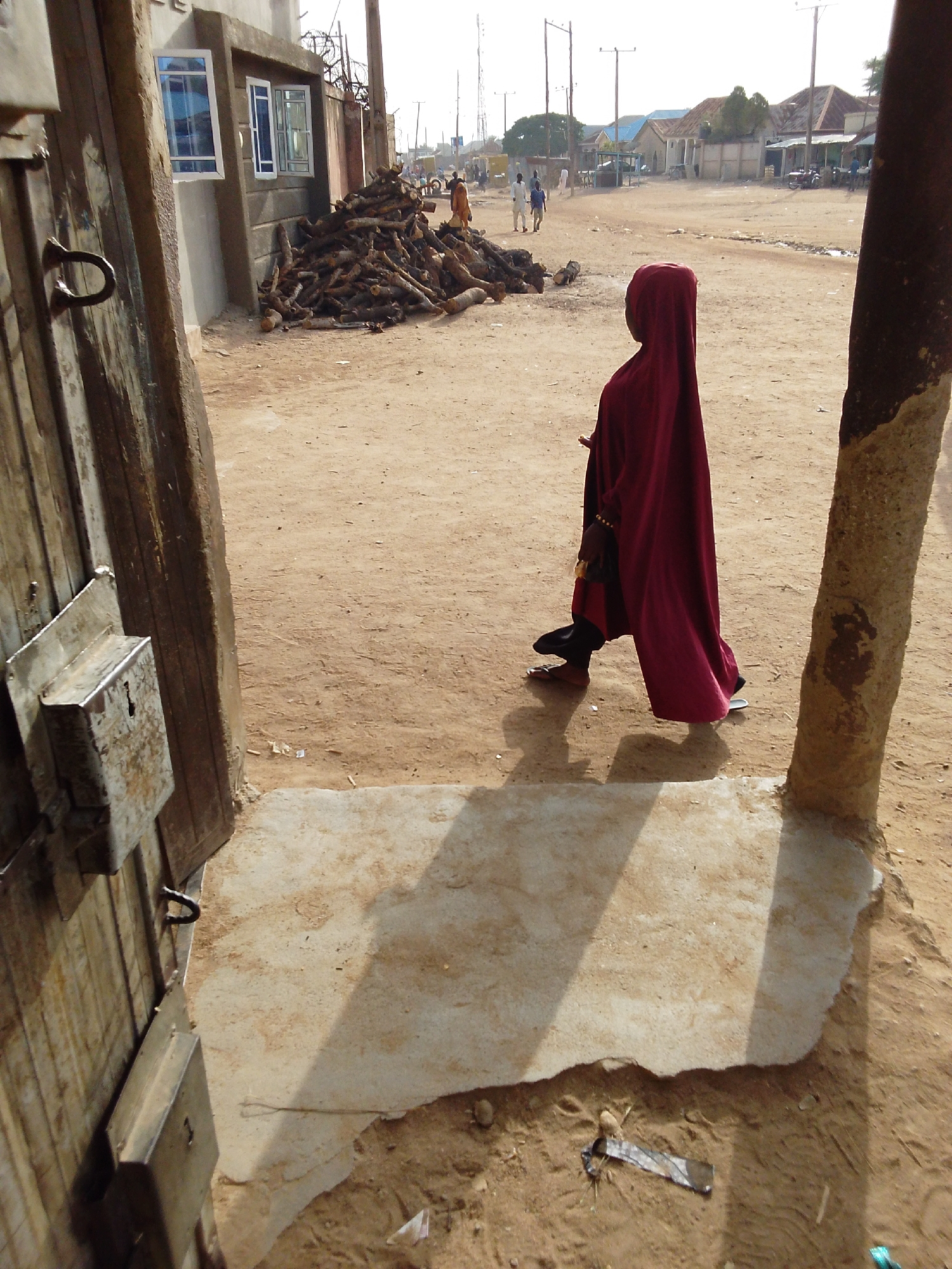 The image shows an outdoor scene in Bauchi, Nigeria. A woman in a long, deep red cloak is walking on a dirt road. She has on black shoes. A pile of stacked wooden logs is visible to her left, near a building with windows. People walk in the distance. An open wooden gate is in the foreground on the left, while a weathered pole is on the right. The ground is predominantly dirt. There is trash near the base of the front door frame.