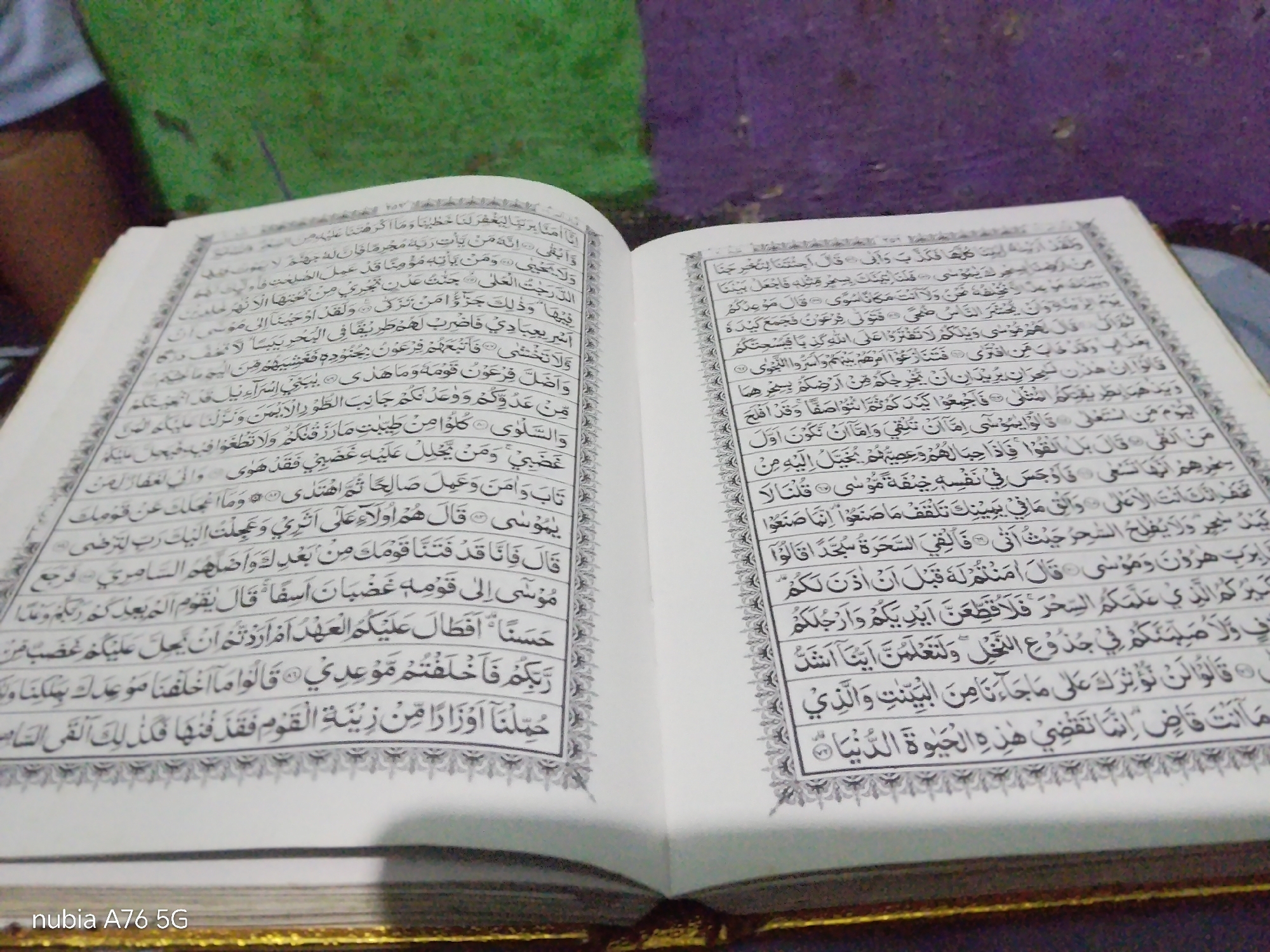 An open Quran rests on a dark surface, likely a table or floor, with a glimpse of purple and green fabric in the background. The Quran is open to a spread of Arabic text, predominantly black ink on white pages, with decorative borders. The left page features smaller text blocks and verse numbers, while the right page displays a larger block of text. The lighting suggests an indoor setting, possibly with ambient light contributing to the scene. The image appears to be taken from a slightly elevated angle, looking down at the book. The bottom left corner of the image shows a watermark: "nubia A76 5G".