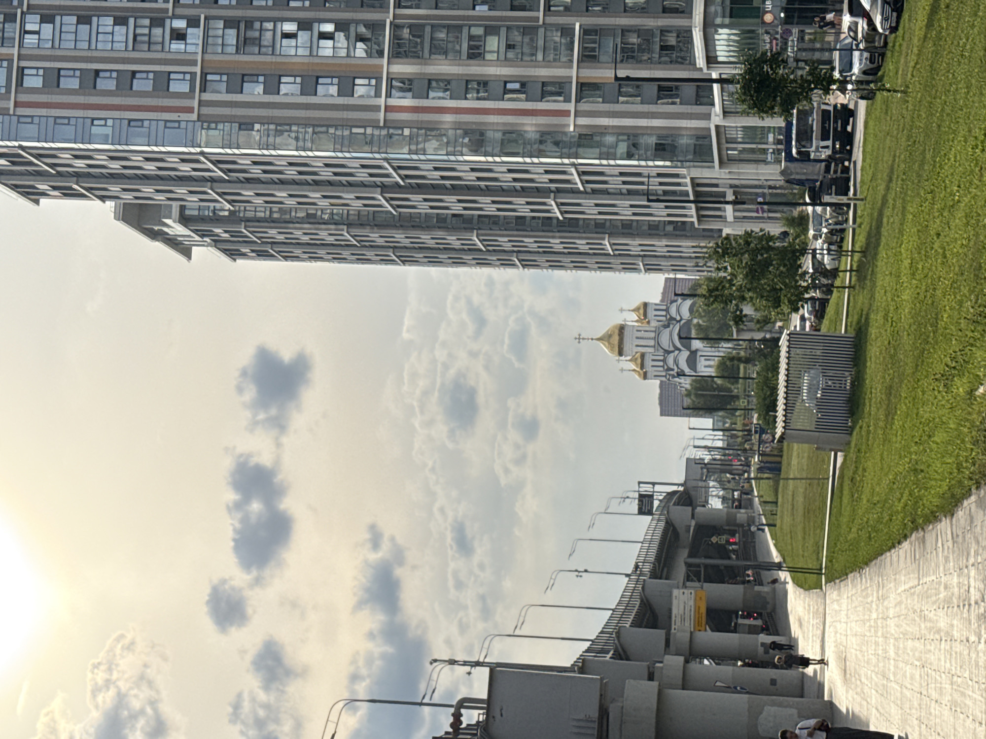 The image depicts a street scene in Moscow, Russia, with a prominent modern high-rise building on the right and a large bridge structure on the left. In the background, a white cathedral with golden domes is visible. The sky is partly cloudy with the sun shining through, casting a bright light.
On the left side of the image, the elevated bridge structure is supported by large concrete pillars. Streetlights line the bridge. A paved pedestrian walkway runs alongside the bridge, with a grassy area to the right of the walkway. Several people are walking on the path.
The tall building on the right is a modern residential or office complex, characterized by numerous windows and a contemporary architectural design. Its facade features a mix of glass and grey panels, with vertical red accents.
Further down the street, a line of cars is parked on the right side of the road. A garbage truck is also present. In the distance, the white cathedral with its multiple golden domes stands out against the sky. The overall scene suggests an urban environment in Moscow, blending modern architecture with religious landmarks.