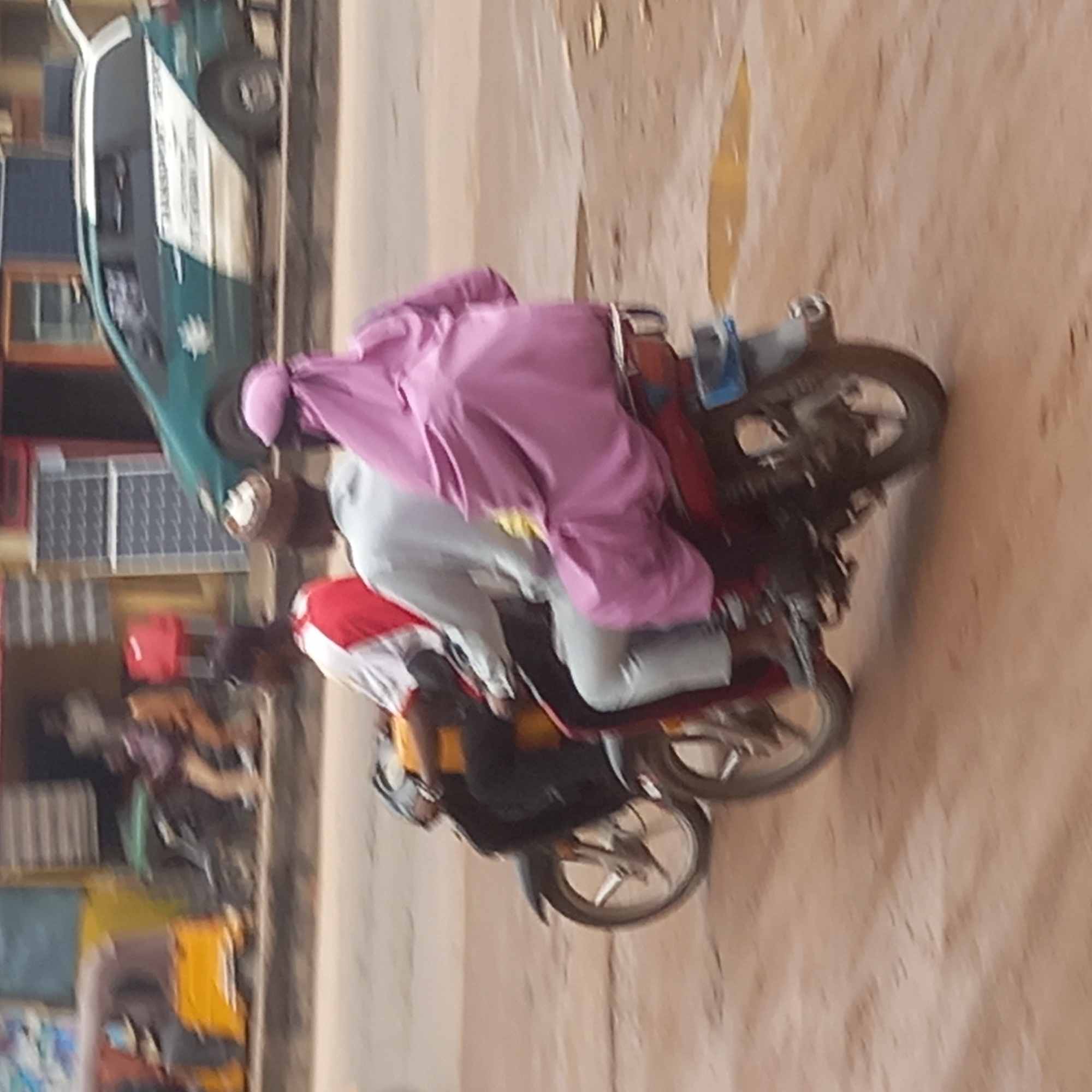 In Dadi Riba, Nigeria, a photograph shows a motorcyclist driving on a paved road, accompanied by a passenger. The motorcyclist wears a red-and-white shirt. The passenger is draped in a pink garment. A green car is parked in the background near some buildings.