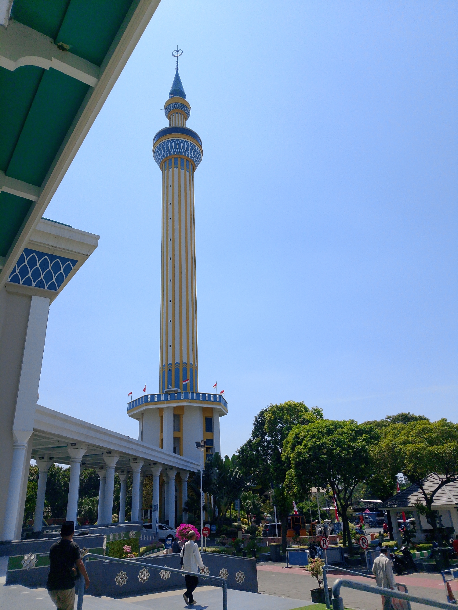 In Surabaya, Indonesia, a tall, slender minaret with yellow and white stripes and blue accents stands against a clear blue sky. The minaret is topped with a blue spire and a crescent moon. In the foreground, a white building with a green roof and blue trim extends from the left side of the frame. A white colonnade connects this building to a walkway. Two men are walking up a set of stairs in front of the walkway. The man on the left wears a black shirt, a traditional Indonesian hat (peci), and khaki pants. The man on the right wears a white long-sleeved shirt and light-colored pants, carrying a pink bouquet. The walkway leads towards the base of the minaret, which has a platform with small flags. Lush green trees and more buildings are visible in the background, along with parked vehicles. The scene suggests a sunny day at a religious or cultural site.