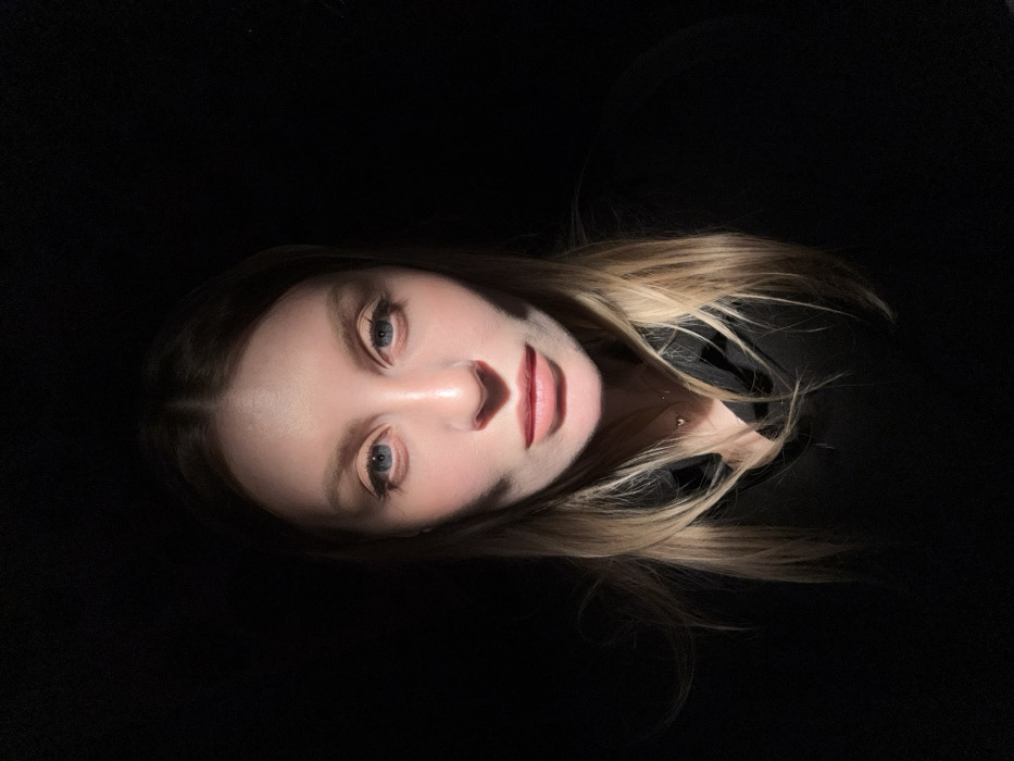 A close-up portrait depicts a single female subject, centrally framed from the chest up, against an entirely dark, indistinguishable background. The individual has fair skin, light blue eyes, and long, straight blonde hair parted in the middle. She looks directly at the camera with a neutral expression, lips closed. A strong, directional light source illuminates her face and upper chest, creating pronounced shadows on the sides of her face, beneath her nose, and under her chin, contrasting sharply with the deep black surrounding areas. She is wearing a dark-colored, possibly black, hooded garment. A delicate, gold-colored necklace with a small heart-shaped pendant is visible around her neck. No specific environmental features or objects are discernible to determine a precise location, therefore, a location in the city of Moscow, Russia, cannot be identified from the visual evidence presented.