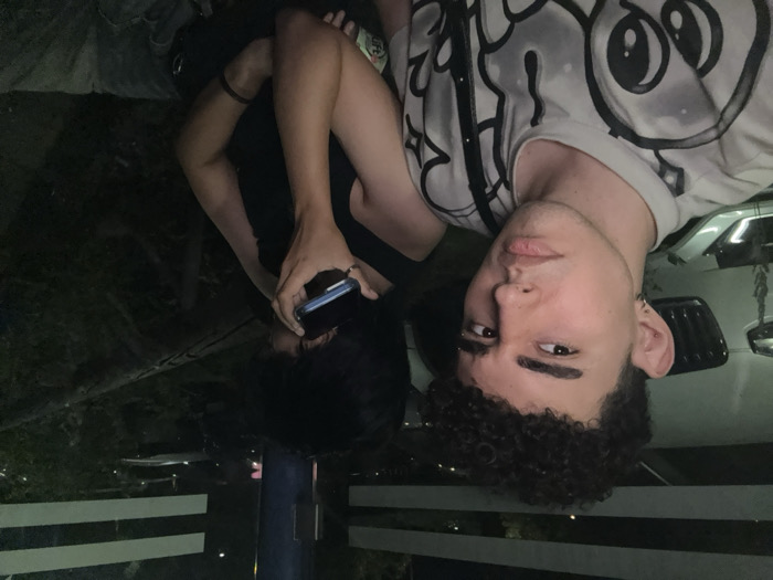 Two people pose closely for a photo. One wears a white t-shirt with a black graphic print and has curly hair. The other wears a black top and holds a phone, partially obscuring their face. They appear to be outdoors at night, with glimpses of a car and vegetation in the background. The image is rotated, giving a slightly disorienting perspective.
