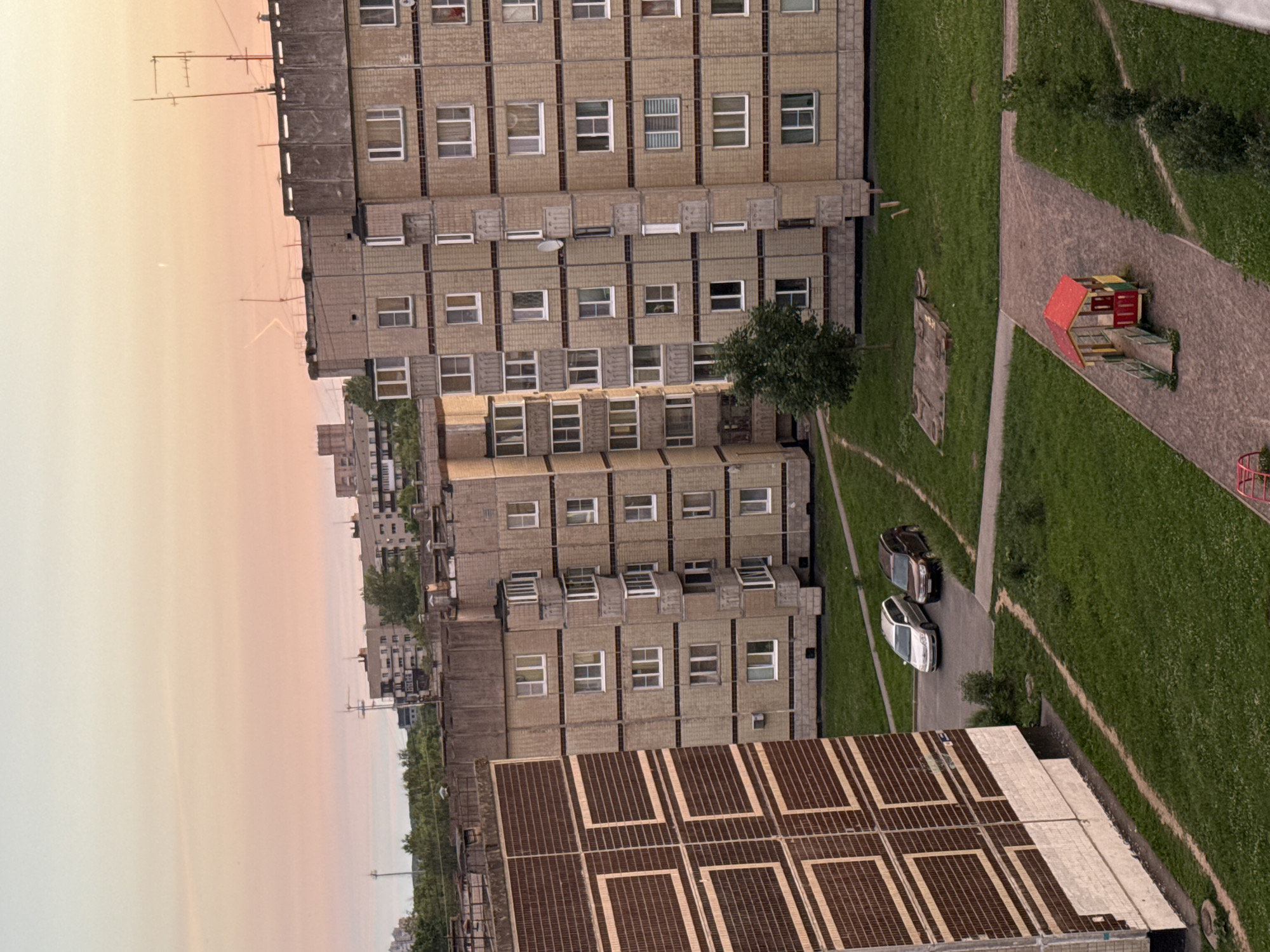 This is an aerial, long-shot, eye-level view of a residential courtyard in Kirishi, Russia, during what appears to be sunset. The sky is a soft gradient of pale yellow and pink hues. Dominating the foreground is a multi-story, beige brick apartment building with numerous windows and balconies. To the left, a section of another apartment building with a patterned brown and beige facade is visible.
In the courtyard, two cars are parked on a paved area: a silver station wagon and a dark brown SUV. A small, red-roofed playhouse with yellow and green accents sits on a paved pathway in the lower right quadrant of the image, accompanied by a small slide structure. A portion of a sandbox is also visible on the grass. Green grass covers the majority of the courtyard area. A few trees are scattered throughout the scene, including a young deciduous tree near the center of the image. The background features more apartment buildings and trees, with visible utility poles and antennas on the rooftops.