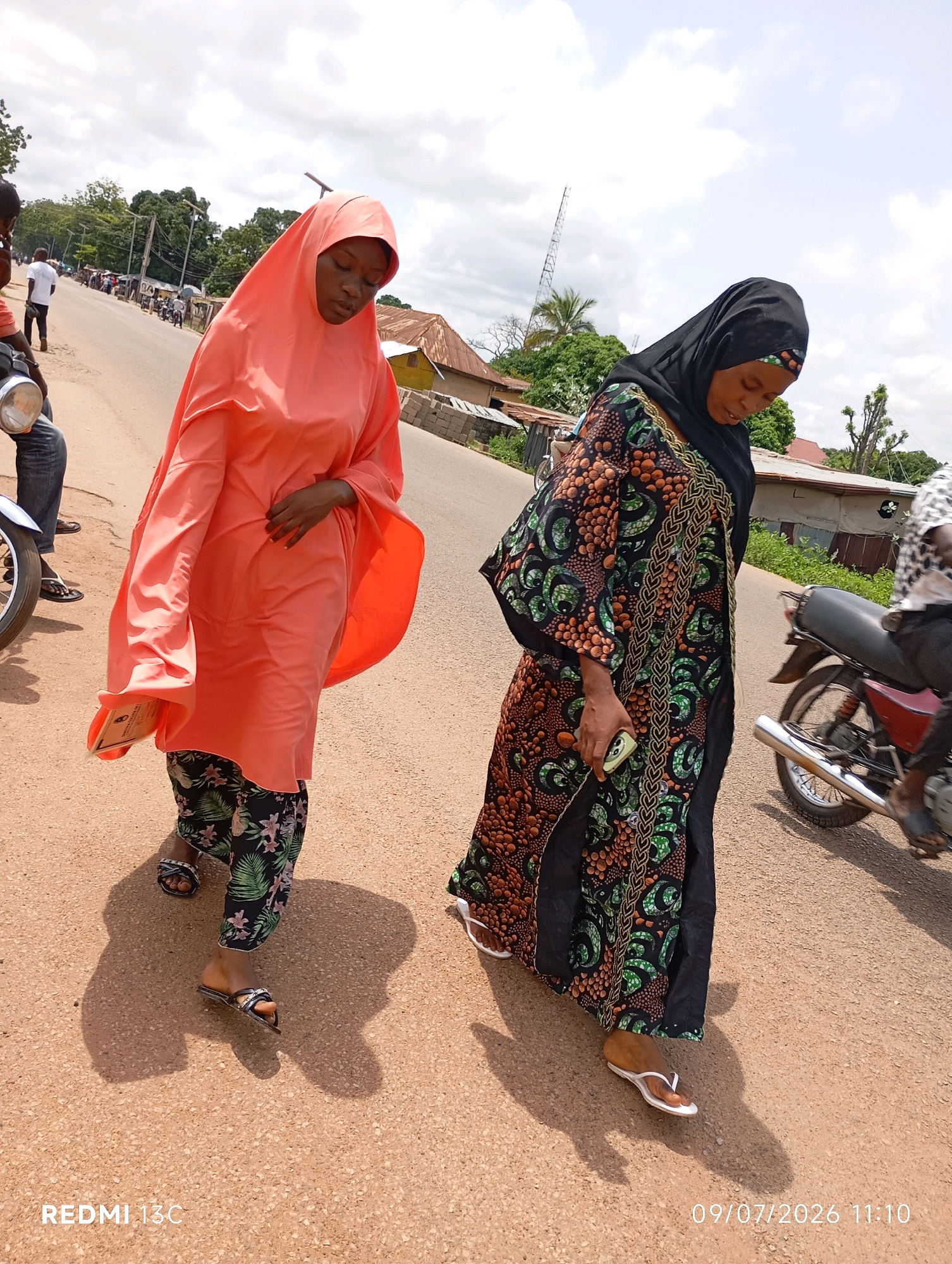 Two women walk on a paved road in Shendam, Nigeria. The woman on the left wears a bright orange hijab and a floral patterned skirt. She holds a folded document in her left hand. The woman on the right wears a black hijab and a black and green patterned dress. She is holding a smartphone in her right hand. A motorcycle is visible in the background to the right, with its rider obscured. The environment appears to be a semi-urban area with buildings and trees lining the road. The sky is cloudy. The timestamp "09/07/2026 11:10" and "REDMI 13C" are visible in the bottom right and bottom left corners respectively.