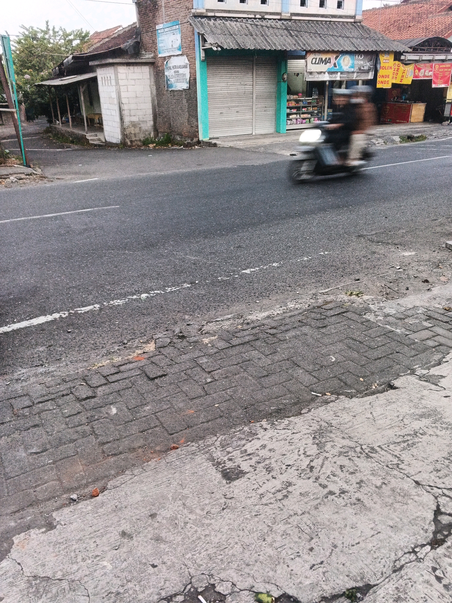 The image shows a street scene in Ungaran, Indonesia. The foreground features a weathered concrete and brick sidewalk next to a paved road. In the mid-ground, a person is riding a motorcycle, blurred due to motion, on the asphalt road. The background consists of a street with several small shops. One shop has a green and white awning, closed metal shutters, and displays various products on shelves visible through a glass section. Another shop is partially visible with a red exterior. Above the shops, the sky is overcast. The general environment suggests an urban or semi-urban setting with commercial activity.