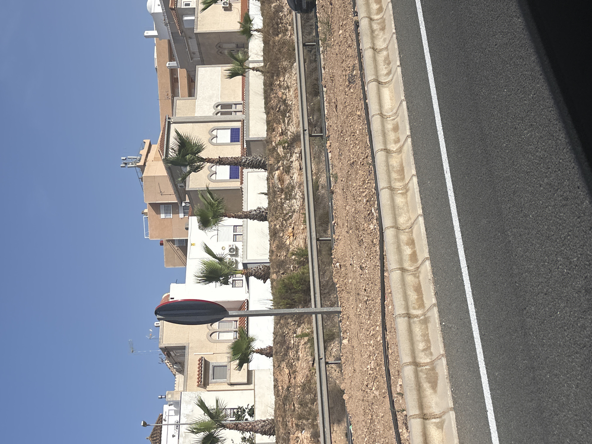 A wide, low-angle outdoor shot shows a row of light-colored apartment buildings with red-tiled roofs and palm trees in front of them, set against a clear blue sky. The buildings are three stories tall with white balconies and arched windows. A traffic barrier with a red and black circular sign is in the foreground, and a paved road with a white line is visible at the bottom of the frame. The ground is covered in dry grass and rocks, with a concrete curb. A black car is partially visible on the right side of the image.