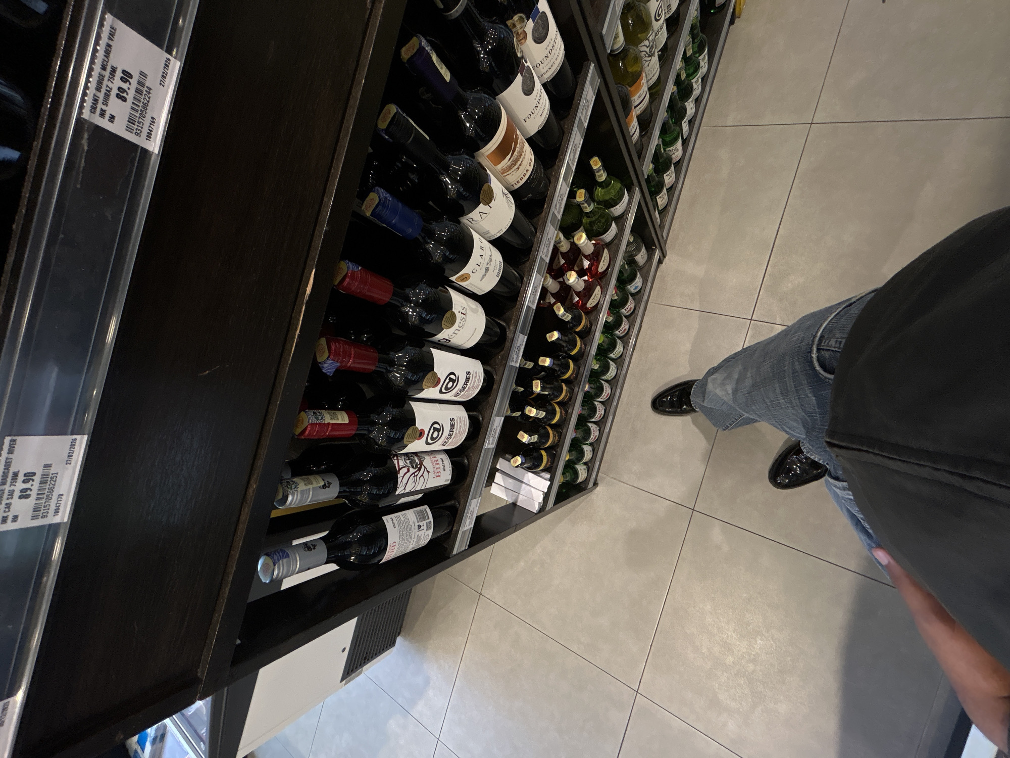 The image depicts a person standing in front of a wine display in a retail store. The person is wearing jeans and black shoes. The wine bottles are arranged on shelves. The bottles are of various brands and types. The store appears to be located in Subang Jaya City Council, Malaysia, indicated by the price labels on the display cases with prices in RM. The price label on the left is for "INK CAB SAU 750ML MARGARET RIVER" for RM89.90, and the one on the right is for "GRANT BURGE MCLAREN VALE INK SHIRAZ 750ML" for RM89.90. The "27/02/2026" is also printed on the labels, next to the barcodes. The floor is tiled.