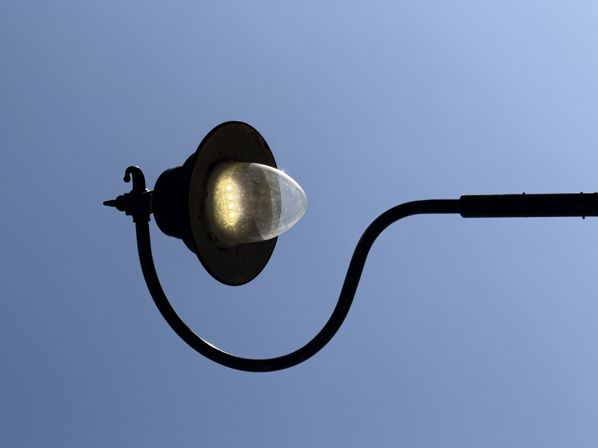 A low-angle, daytime shot shows a black street lamp against a blue sky. The lamp has a rounded, transparent cover that appears to be illuminated. The lamp fixture is attached to a curved arm that extends horizontally to the right. The sky is uniformly blue without any clouds.