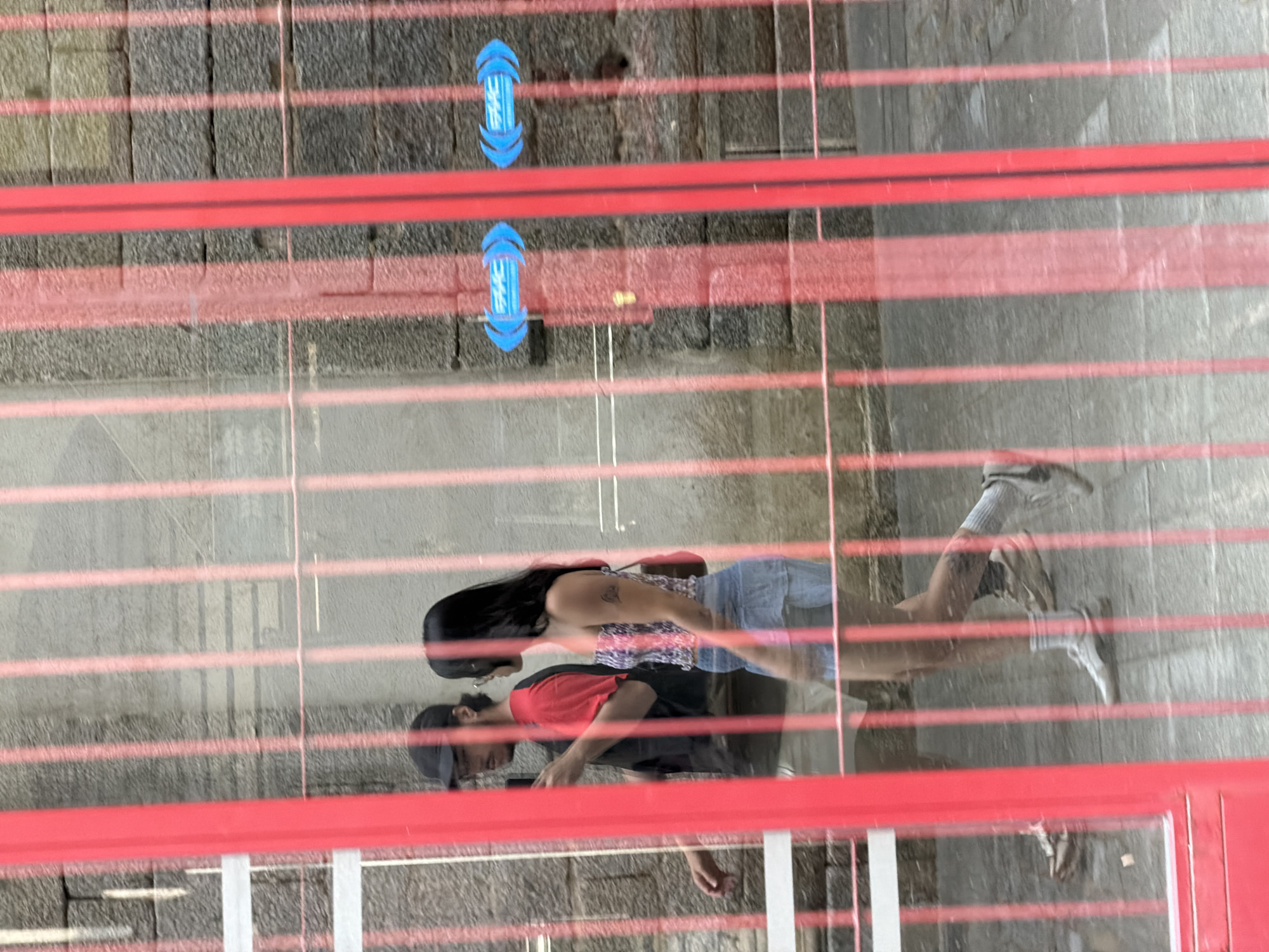 This media file shows a reflection on a glass surface, viewed from above. The subjects are two people, appearing as a couple, with one person wearing a red and black shirt and a baseball cap, and the other wearing a patterned top and denim shorts. They are walking together, with their reflection showing them facing each other slightly. The scene appears to be an outdoor area, possibly a pathway or a plaza, with a stone or concrete ground. Red horizontal bars are prominent in the foreground and mid-ground, creating a striped effect across the image, and are likely part of a railing or barrier. Two blue directional arrows with text are visible on the ground, indicating movement or guidance. The lighting suggests it is daytime. The location is identified as San Lorenzo de El Escorial, Spain. There are no visible emotions, specific weather conditions, or time of day cues beyond daylight. The text on the blue arrows is illegible.