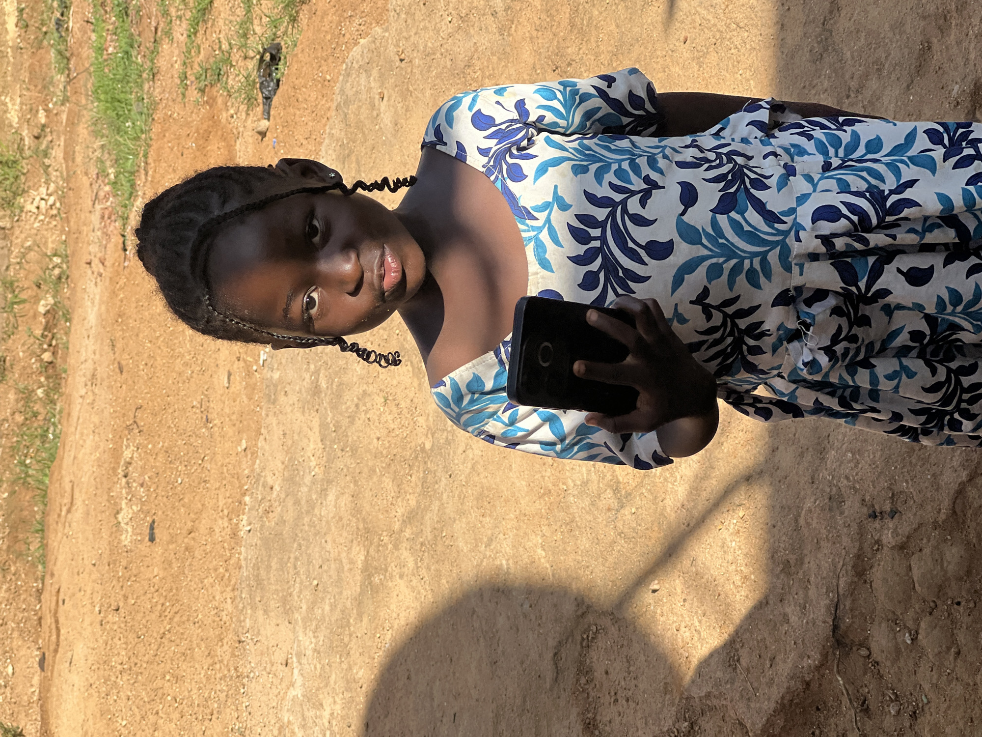 A young girl, with dark skin and braided hair styled in intricate twists, stands outdoors, holding a black smartphone. She is wearing a white dress adorned with a repeating pattern of blue leaves and branches. Her gaze is directed towards the viewer, with a neutral expression. The background consists of a dusty, reddish-brown ground and some sparse green grass. A large, soft-edged shadow, cast by an unseen object, covers the left side of the frame, indicating sunlight. The overall lighting suggests a bright, sunny day. The environment appears to be an outdoor area within Jos, Nigeria, possibly a residential compound or a less developed urban space, given the unpaved ground and natural elements.