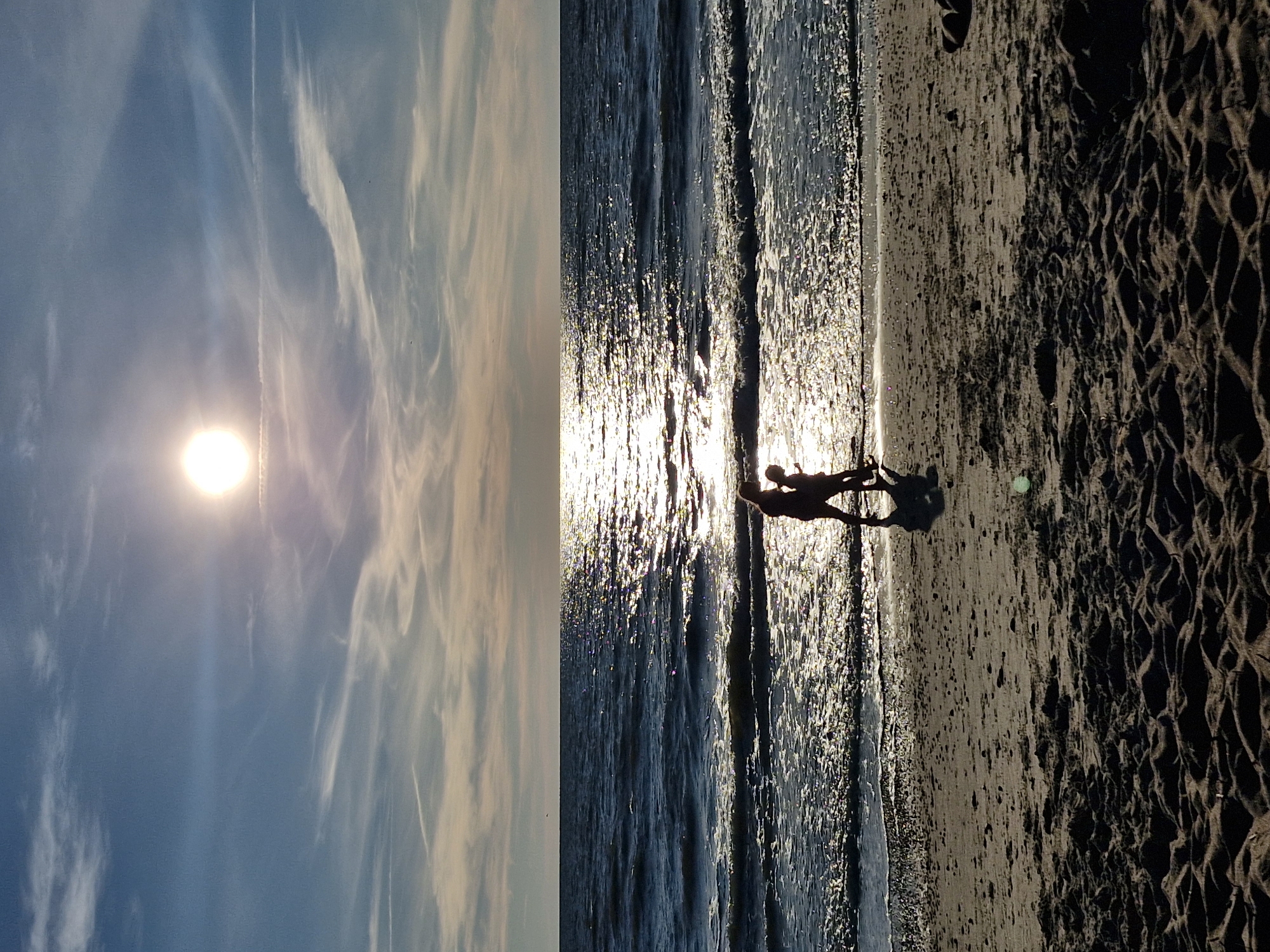 This is a vertical, full-shot, color photograph taken from a low angle of a beach scene at sunset. The sun, a bright orb, is high in the sky, casting a shimmering reflection across the dark blue water. The water has gentle waves breaking on the shore. Two silhouetted figures, appearing to be adults or older children, stand close together on the wet sand at the water's edge, facing the sun. Their shadows are cast long on the beach. The sand in the foreground is dark and textured, with ripples and indentations. The sky above is a gradient of blue and light orange, with wispy white clouds. The scene is indicative of a coastal environment, likely the beach in Karklė, Lithuania, given the typical sunset lighting and landscape.