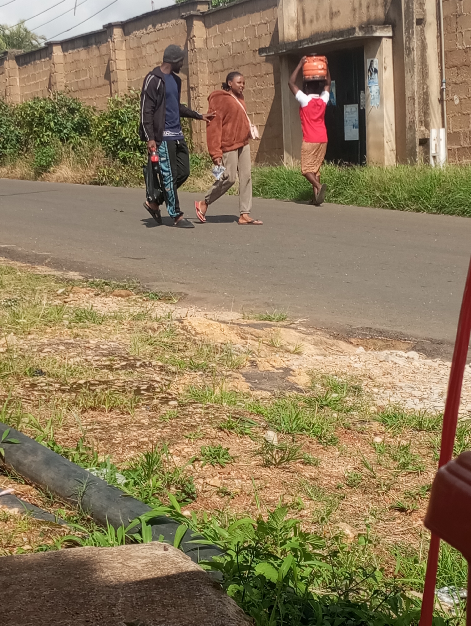 In Jos, Nigeria, three individuals are walking on a paved road. A male on the left, wearing a dark beanie, a dark jacket with white stripes, and dark pants with blue stripes, carries a red and black bottle in his right hand. He is walking next to a female wearing a brown hooded jacket and light-colored pants. On the right, a female walks with an orange container balanced on her head, wearing a red t-shirt and a patterned skirt. The environment features a brick wall on the left, overgrown green grass, and a building with a tan-colored facade on the right. The road appears worn with some patches of exposed dirt and gravel. In the foreground, a black pipe and a large brown object are visible amongst the greenery.