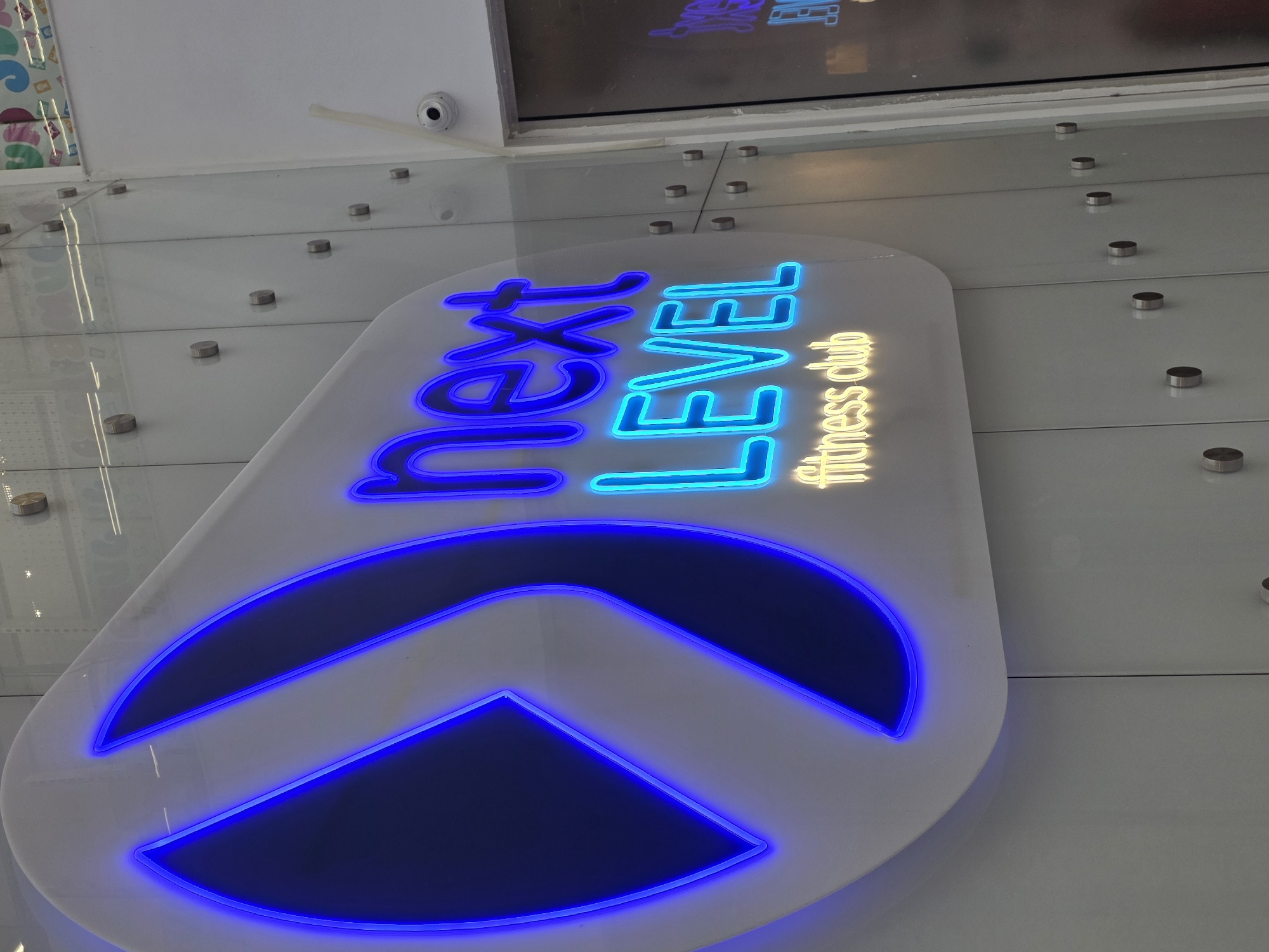 The image shows a sign for "Next Level fitness club" mounted on a wall. The sign is white with blue neon lettering and a blue neon arrow pointing upwards. The words "fitness club" are smaller and in yellow neon. The wall appears to be part of a building interior. The location is Plovdiv, Bulgaria. There are small metal discs attaching the sign to the wall. A camera is visible near the top of the frame. The image appears to be taken from below, looking upwards at the sign.
