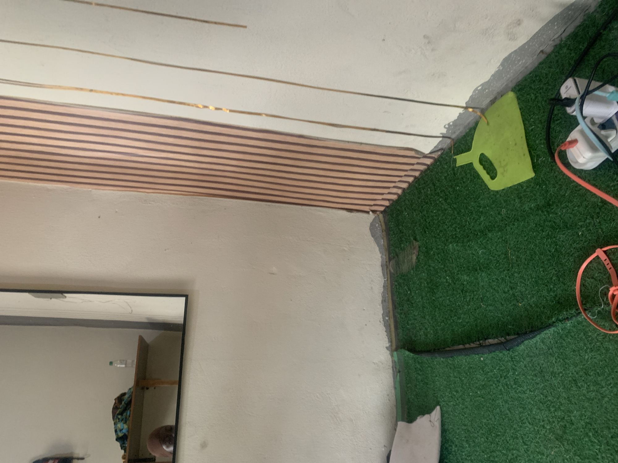 The image depicts an indoor scene, possibly a room, with a green, artificial turf-like floor covering. A grey, peeling edge is visible where the floor meets the wall in the foreground. A bright green dustpan lies on the turf near a wall adorned with vertical, light brown and dark brown striped paneling. To the left of the paneling, a white wall is visible. In the lower right quadrant, a power strip with several plugs and cables is situated on the turf. A portion of a white object, possibly a piece of furniture or bedding, is seen on the far left edge of the frame, also on the green floor. In the background, a mirror reflects a wooden shelf with items, including a bottle of water and some fabric. A person's head, wearing a brown headscarf, is partially visible in the reflection of the mirror, looking downwards. The lighting suggests an indoor environment, possibly with artificial light. The overall impression is one of a somewhat unkempt or under-renovation space. No specific location in Jauro Yinu, Nigeria is identifiable from the visual cues within the image itself.