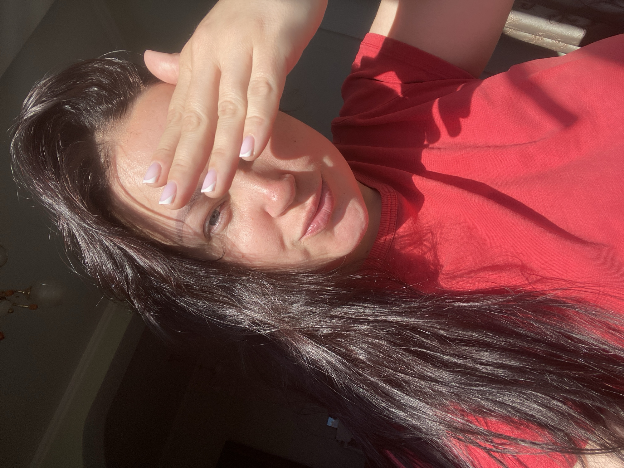 A selfie taken from a low angle depicts a light-skinned person shielding their face from direct sunlight with their right hand. The hand has nails painted with a white French tip. The person has dark hair and is wearing a red t-shirt. The background is partially obscured by shadow, but reveals a glimpse of a wall and potentially a window. The image contains no definitive indicators of the city of Sochi, Russia.