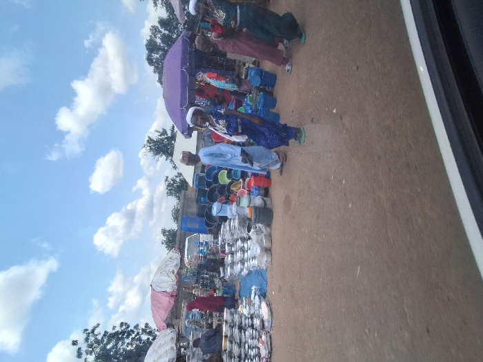 The scene depicts an active outdoor market situated on unpaved reddish-brown ground in Kungana, Nigeria, under a bright blue sky with scattered white cumulus clouds.
In the left foreground, the white frame of a vehicle is visible, indicating the vantage point. The market comprises multiple stalls, two of which are prominent: one under a light-colored umbrella (white or very pale pink) on the left, and another under a dark purple umbrella on the right.
The left stall features numerous stacks of various sizes of metal pots, bowls, and plates, likely aluminum or stainless steel, arranged directly on blue fabric and the ground. A person wearing a red garment and head covering is partially visible behind this stall. Further back, a white van is parked.
Towards the center and right, extensive displays of assorted plastic buckets and containers in colors including black, red, blue, and green are arranged on the ground.
Several individuals are present, dressed in traditional attire. In the mid-ground, a male wearing a light blue garment and a light-colored cap walks towards the right. To his left, a female in a dark blue patterned dress, white head covering, and light green footwear walks in the same general direction. Another female in a dark green garment and light green footwear is visible walking towards the right side of the frame. Multiple other individuals, predominantly female, wearing various colored dresses and head coverings (including red, maroon, and blue), are standing or moving near the purple umbrella stall and deeper into the market. The overall scene suggests ongoing commerce and pedestrian activity within the market environment.