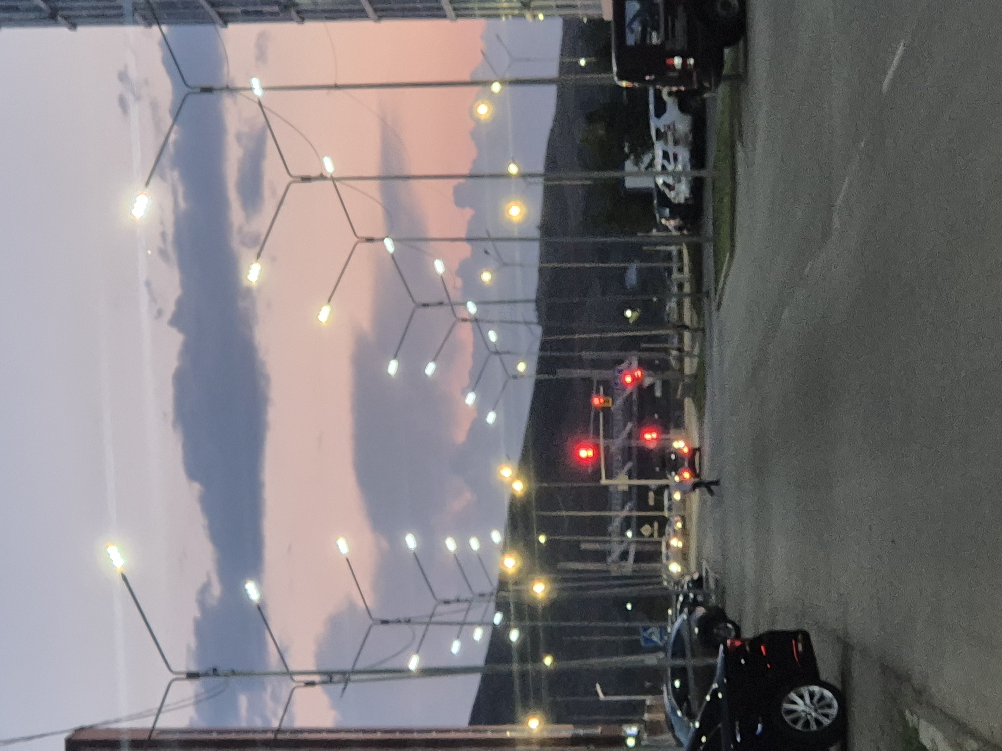 The image depicts a street scene in Kokshetau, Kazakhstan, during twilight. The sky is a blend of pink and purple hues, with dark clouds present. Numerous streetlights are illuminated, casting a yellow and white glow. A building, possibly an apartment complex, is visible on the right side of the frame. The street is paved, with a white line marking the center. Several cars are parked along the side of the road and on the street. A pedestrian is crossing the street in the mid-ground. Traffic lights are visible, with one showing a red light. The overall impression is an urban setting at dusk, with artificial lighting beginning to dominate the fading natural light.
