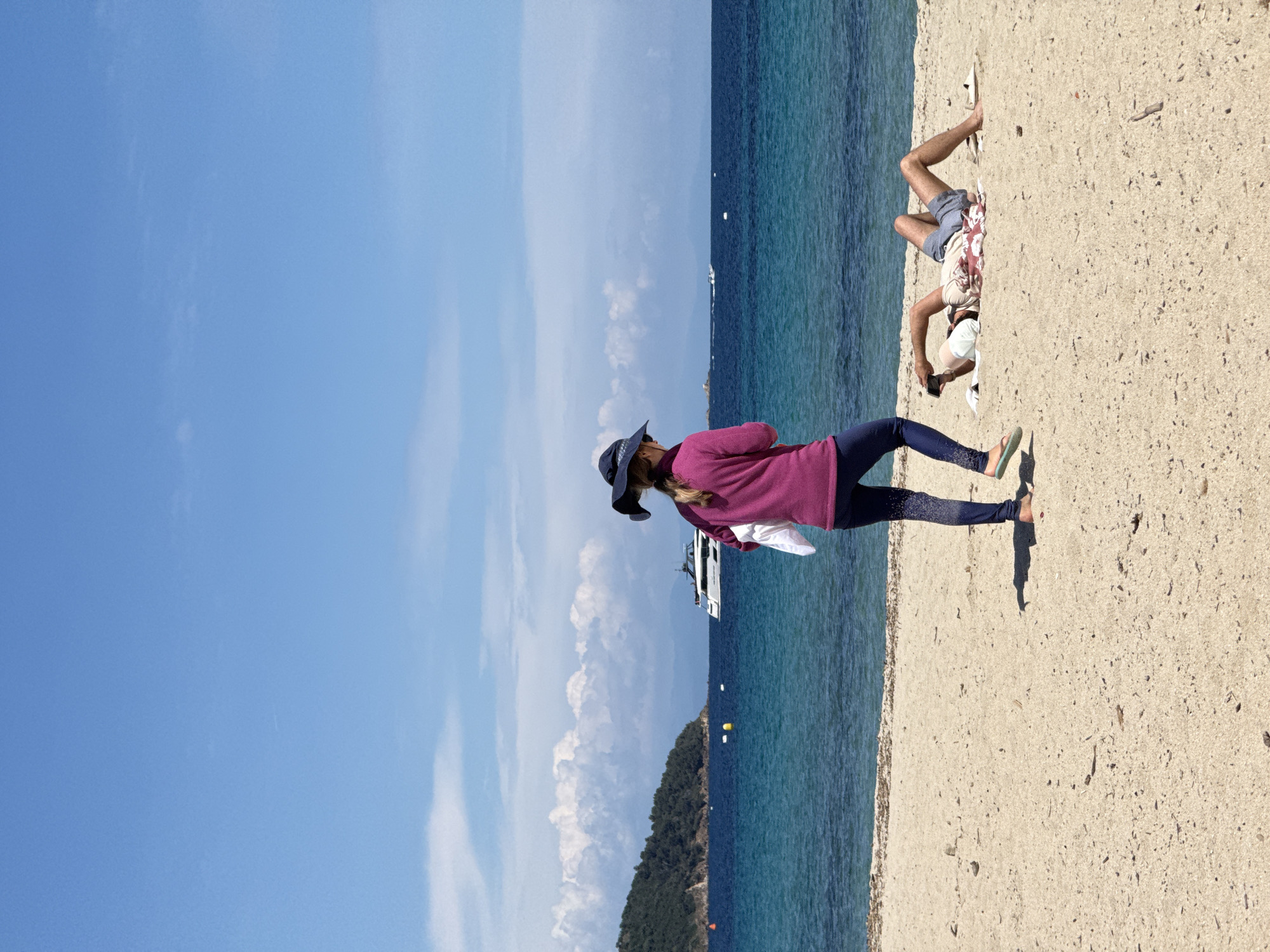 The image shows a beach scene in Ramatuelle, France. A woman wearing a purple long-sleeved shirt, dark leggings, a hat, and sandals stands on the beach. Beside her, a man is lying on the sand, partially obscured. The background features a blue sky, turquoise water, a small island with green foliage, and a yacht.