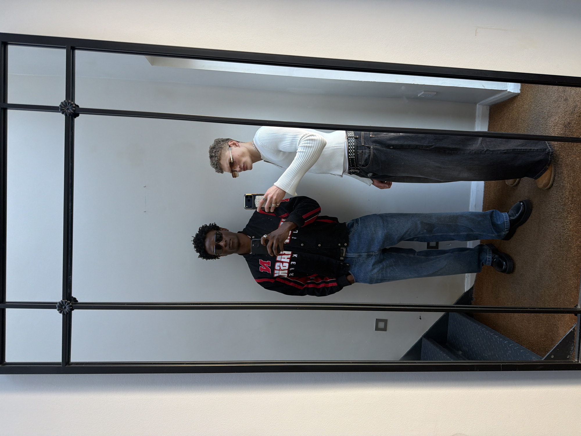 The image shows a mirror reflecting two individuals, presumably taking a selfie. One individual wears a white ribbed shirt, dark jeans with a studded belt, and sunglasses. The other wears a black jacket with red stripes and lettering, blue jeans, and sunglasses. The mirror is framed in black with decorative elements at the intersections of the frame. The background reflected in the mirror shows a white wall, a carpeted floor, and a glimpse of stairs. The location is not discernible from the image.