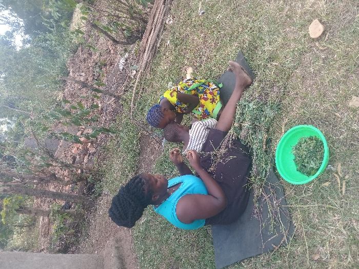 A woman with dark hair sits on a black mat in a grassy area, wearing a blue top and dark skirt. Two children, one in a colorful yellow dress and the other in a striped shirt, sit near her. They seem to be preparing food, as a green bucket filled with greens sits nearby. Debris, twigs, and grass surround them, and trees and vegetation form a backdrop. The scene suggests a rural or outdoor setting.
