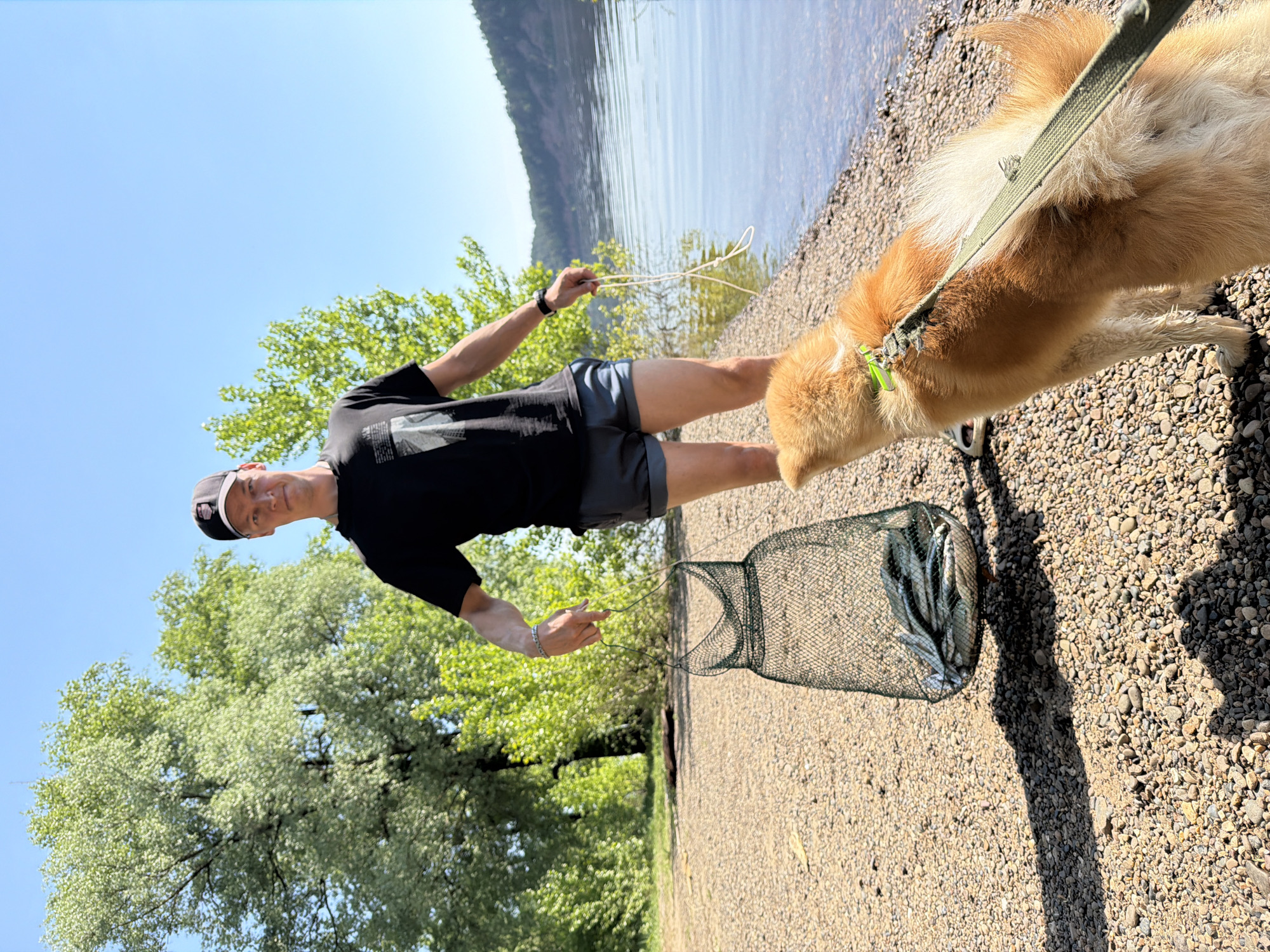 A man is standing on a pebble beach next to a lake with a dog. He is holding a string with a fish net containing several fish. The lake appears to be near some mountains with trees in the background. The location is identified as Ol'khovskoye, Russia.