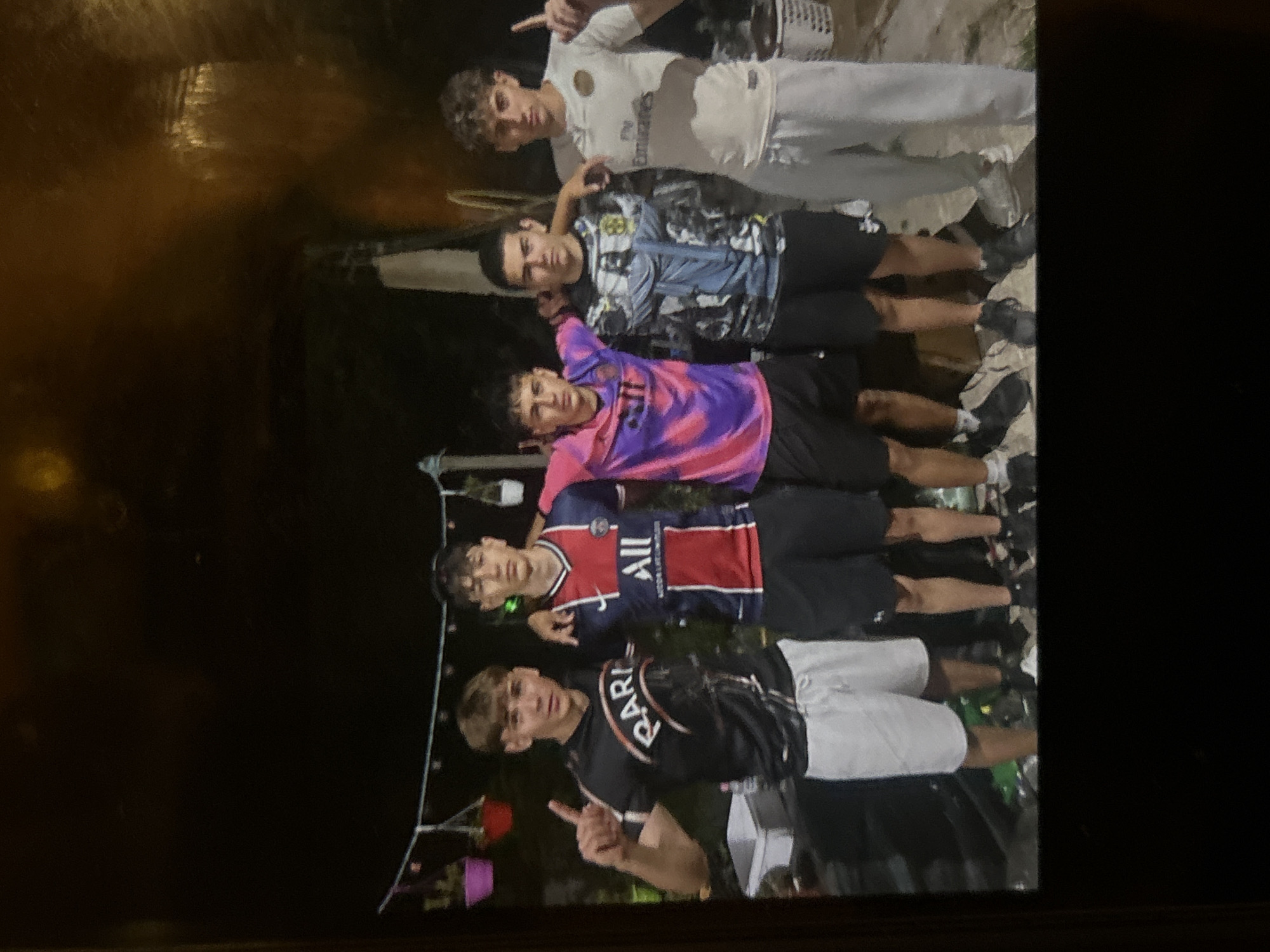 Five young men are posing for a photo outdoors at night, with a dark, possibly wooded, background. They are standing shoulder to shoulder, with their arms around each other. From left to right, the first man is wearing a black t-shirt with "RARI" written on it, and white shorts. He is holding up one finger. The second man is wearing a blue and red striped soccer jersey with "ALL" written on it, and black shorts. The third man is wearing a pink and purple tie-dye t-shirt and black shorts. The fourth man is wearing a t-shirt with an image of Jesus on it and black shorts. The fifth man is wearing a white soccer jersey with "Fly Emirates" written on it, and light gray pants. The overall setting appears to be informal, possibly a backyard party or gathering.