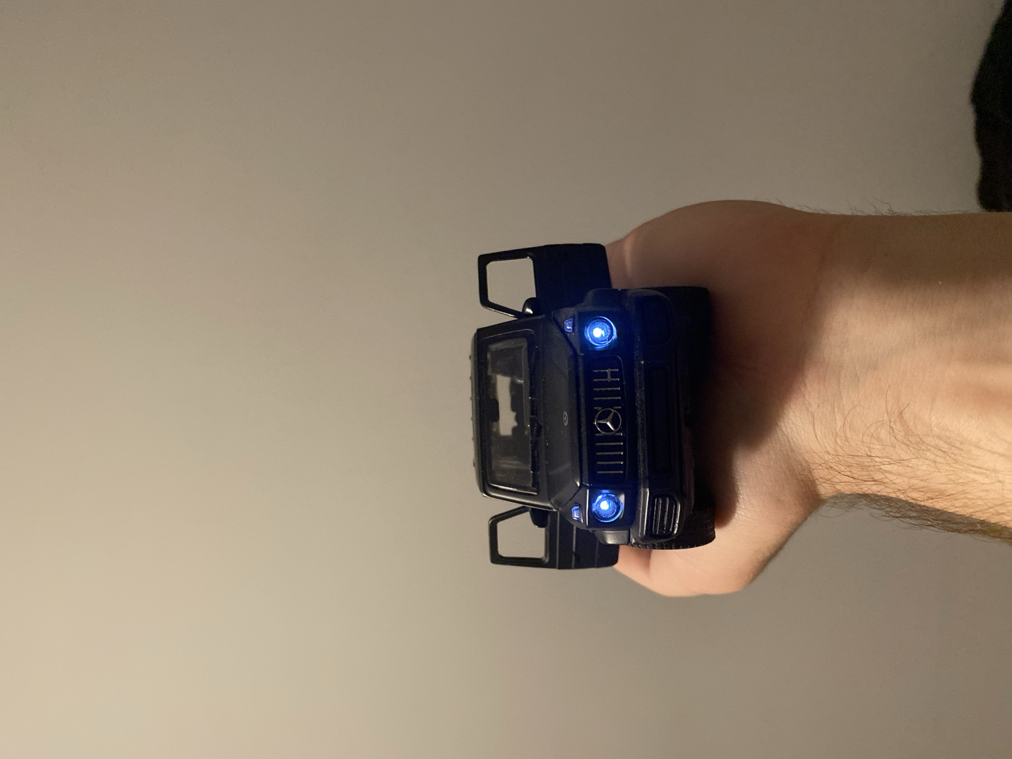 A dark toy Mercedes-Benz G-Class SUV with its doors open and headlights illuminated with blue LEDs is held in a person's hand against a plain, light-colored wall. The car is positioned centrally in the frame. The hand appears to be a man's, with visible arm hair. The background is a neutral, slightly textured wall, suggesting an indoor setting. There are no discernible landmarks or specific environmental features that would indicate a location in Łódź, Poland.