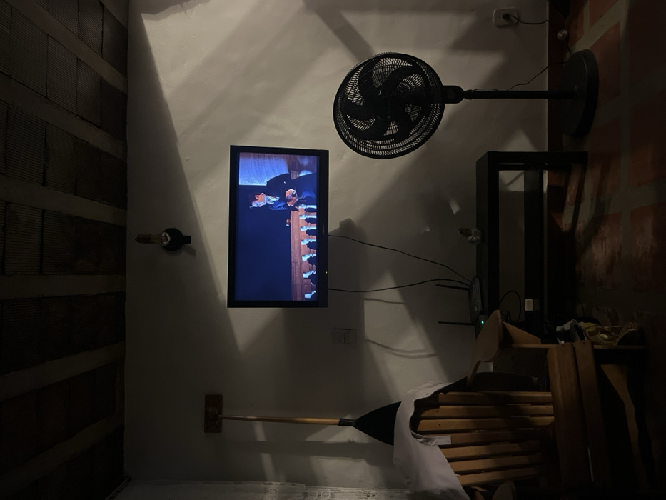A wall-mounted TV displays a scene with a man in a suit. Above it is a black fan. A black metal shelf unit sits to the right, near a brick wall. Below the TV, a paddle is mounted horizontally to the wall, extending over a stack of wooden chairs covered with a white cloth. A wine bottle holder is on the left wall, with brick texture. The room appears to be dimly lit.