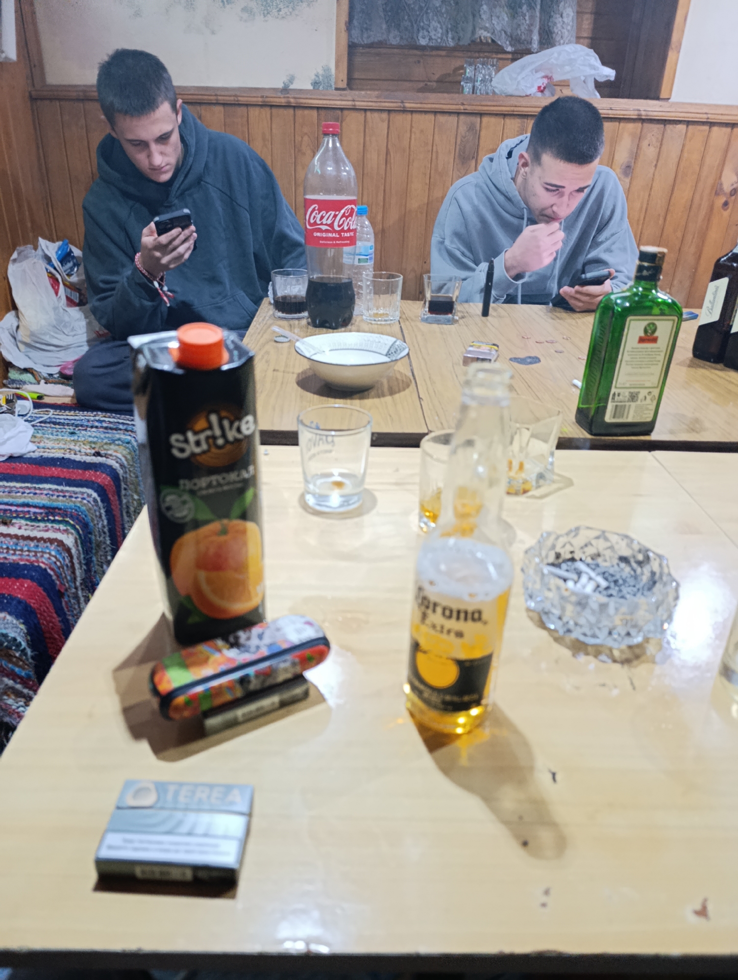 Two young men are seated at a wooden table indoors. Both are looking at their phones. On the table are alcoholic beverages including Coca-Cola, Jagermeister, and Corona beer, along with orange juice, an ashtray filled with cigarette butts, cigarettes, and a lighter. The location is in a room with wood paneling. While Silistra, Bulgaria was requested, it is not possible to confirm this location from the image alone.