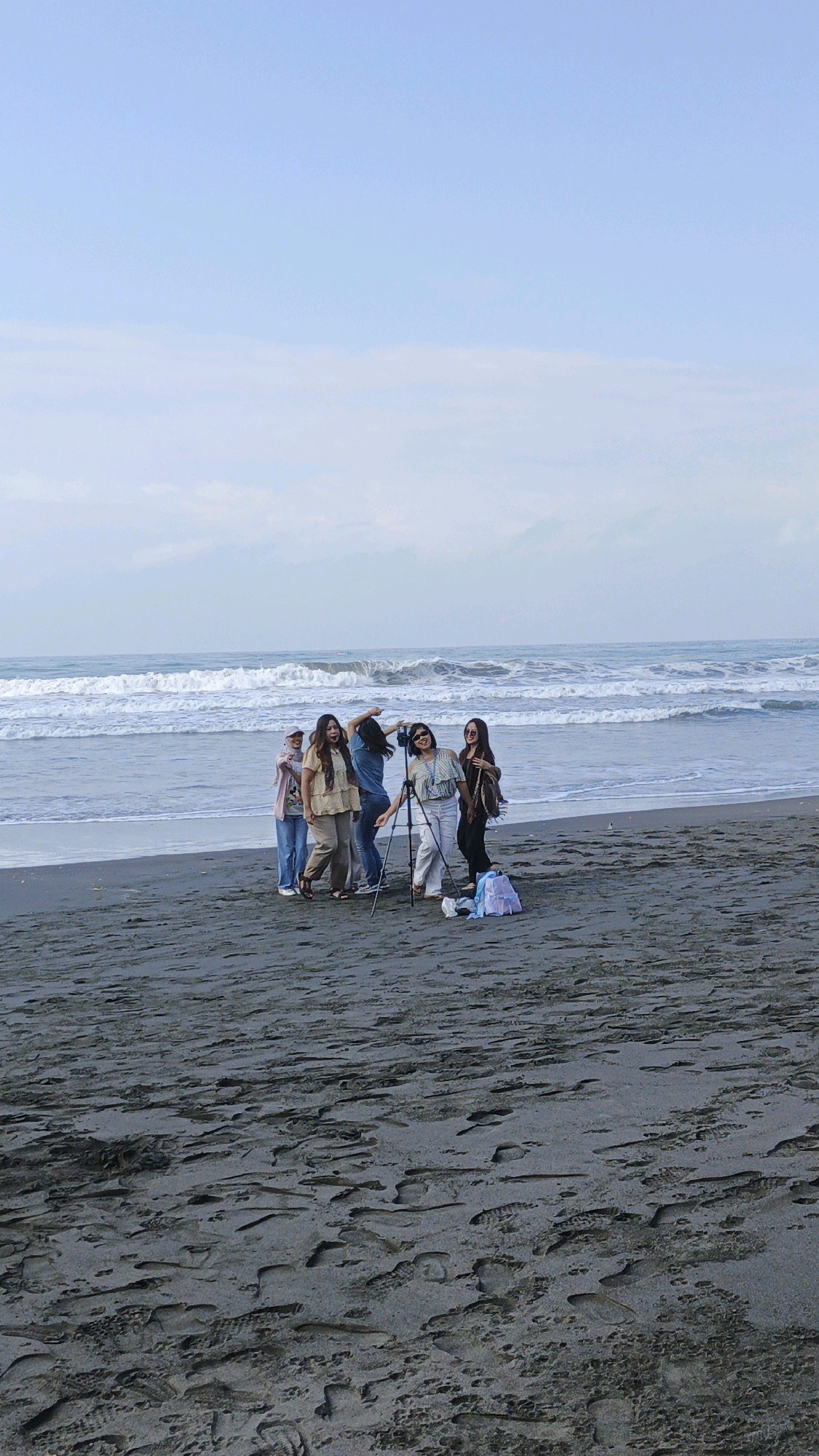 In Desa Ciliang, Indonesia, on a dark sand beach, a group of five women are gathered near the waterline, posing for a photograph being taken by one of them. The ocean, with its gentle waves, serves as the backdrop. The sky is a soft blue with hints of clouds. The sand is covered with numerous footprints. The women are dressed casually, some in light-colored outfits, and one has a camera on a tripod. There is a light-colored backpack on the sand near the group.