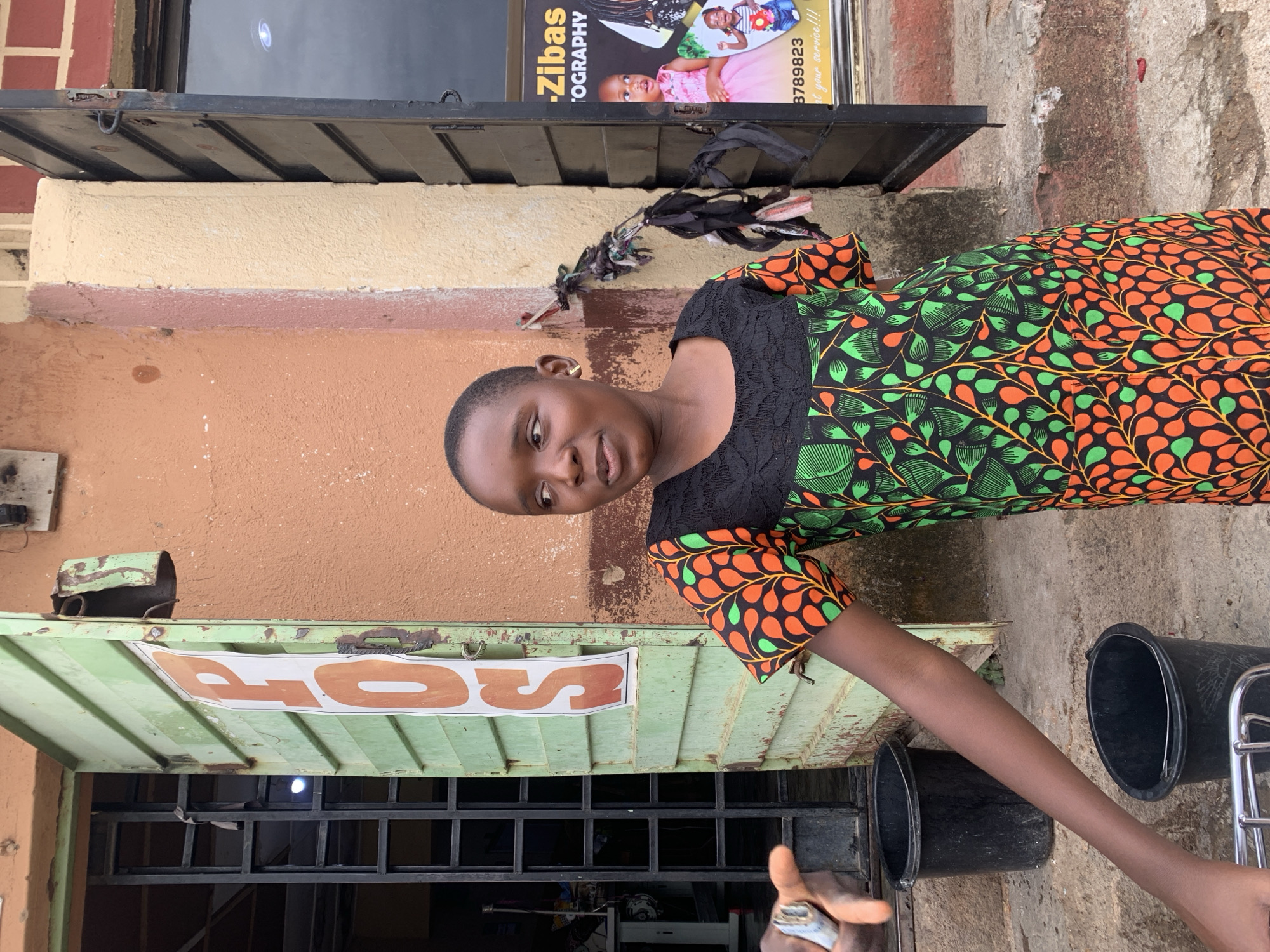 The image displays a young, dark-skinned person standing outdoors, looking directly at the viewer with a neutral expression. She has short, dark hair and wears a small, gold-colored earring in her right ear. Her attire consists of a short-sleeved dress with a black lace-patterned top and a vibrant lower section featuring a striking pattern of green leaves and orange ovals or berries on a black background. Her right arm extends downwards and slightly to her side, while her left arm is mostly obscured.
The setting appears to be a street or commercial area, likely in Jos, Nigeria, in front of a building. To the person's left is a partially open green metal roll-down shutter or door, adorned with orange lettering on a white background that reads "OS." Below the shutter, a black metal security gate is visible. On the ground in front of this area, there are two black plastic buckets and part of a metal rack.
Above and to the right of the person, a photography studio sign is partially visible. The sign features the text "Zibas 'OGRAPHY" (likely part of "Zibas Photography"), a phone number "3789823," and the phrase "...at your service!!!," along with images of young children. The wall behind the person is painted in sections of brown, light tan, and reddish brick at the top. A dark metal awning or roof extends above the sign. The natural, bright lighting indicates it is daytime. A partial view of another person's hand, holding a metallic object, is visible in the bottom right corner.