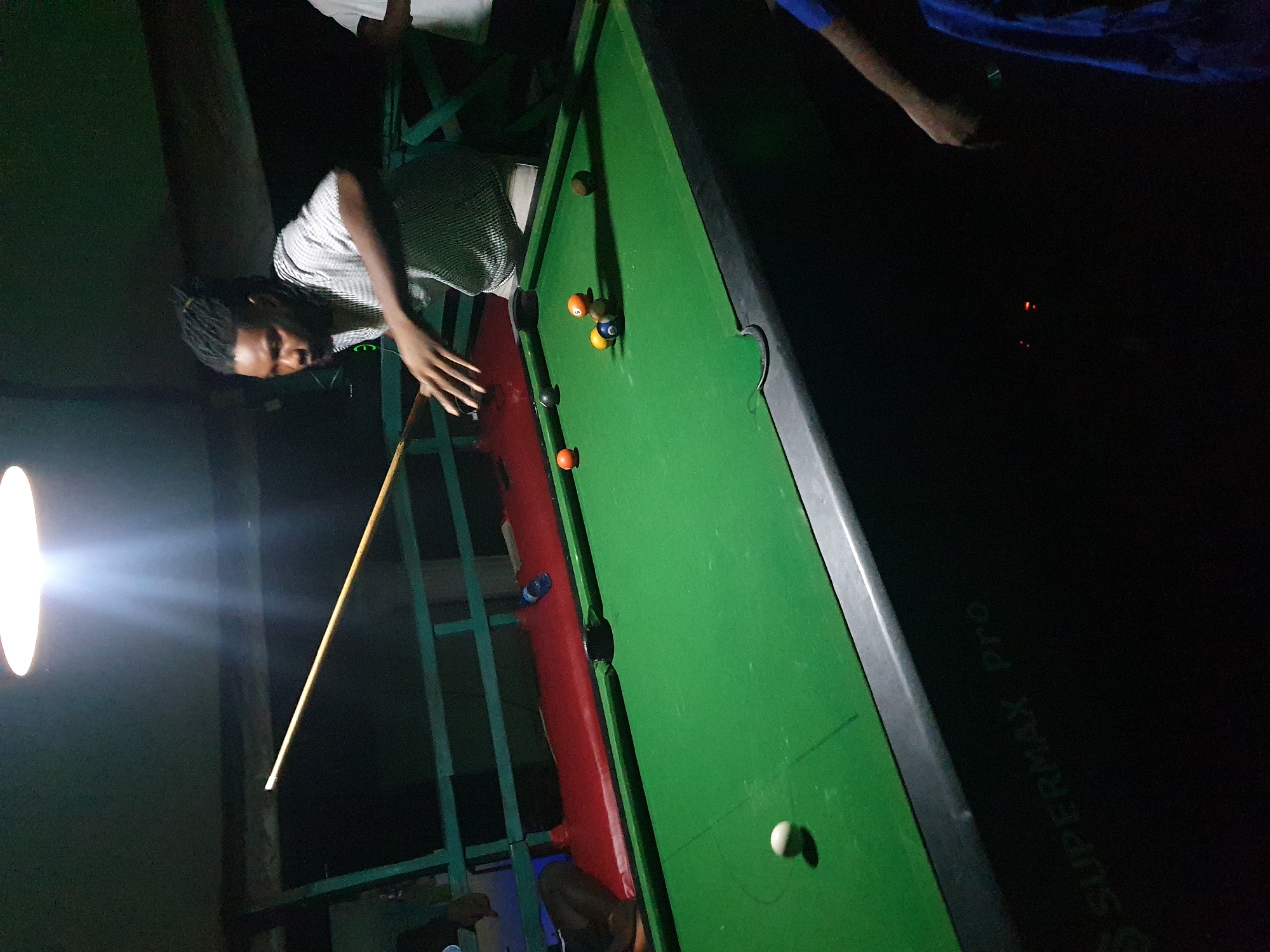 A dimly lit indoor pool hall in Lokoja, Nigeria, features a man with dreadlocks playing pool. He is wearing a striped shirt and is in the process of striking a cue ball with a pool cue. Several billiard balls, including the cue ball, a solid orange ball, a solid black ball, a striped blue and white ball with the number 5, a solid yellow ball, and a solid brown ball, are scattered on the green felt surface of the pool table. The table is branded "SUPERMAX Pro". In the background, a red upholstered bench is visible. Another person, partially obscured, is standing to the right of the pool player. The environment is dark, with a bright overhead light illuminating the playing area.