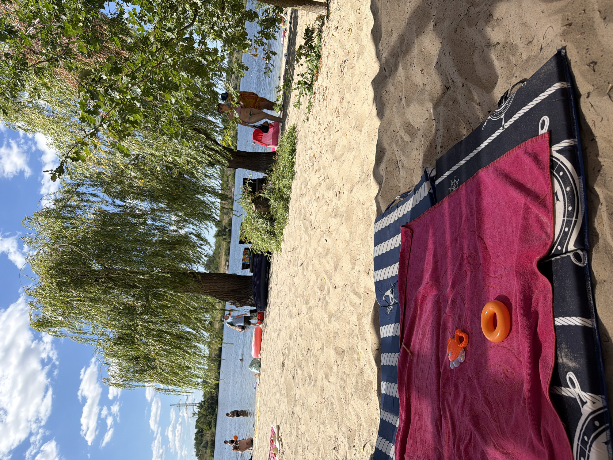 The image depicts a sunny day at a sandy beach on the banks of the Teteriv River in Marianivka, Ukraine. In the foreground, a red towel is spread over a blue and white striped beach mat, with two orange children's toys on the towel. Behind the towel, people are visible in the shallow water and on the shore. Several large willow trees provide shade on the sandy bank. The Teteriv River flows calmly, with a few people swimming or wading in the water. In the distance, across the river, the opposite bank is lined with trees and some distant structures. The sky is blue with scattered white clouds.