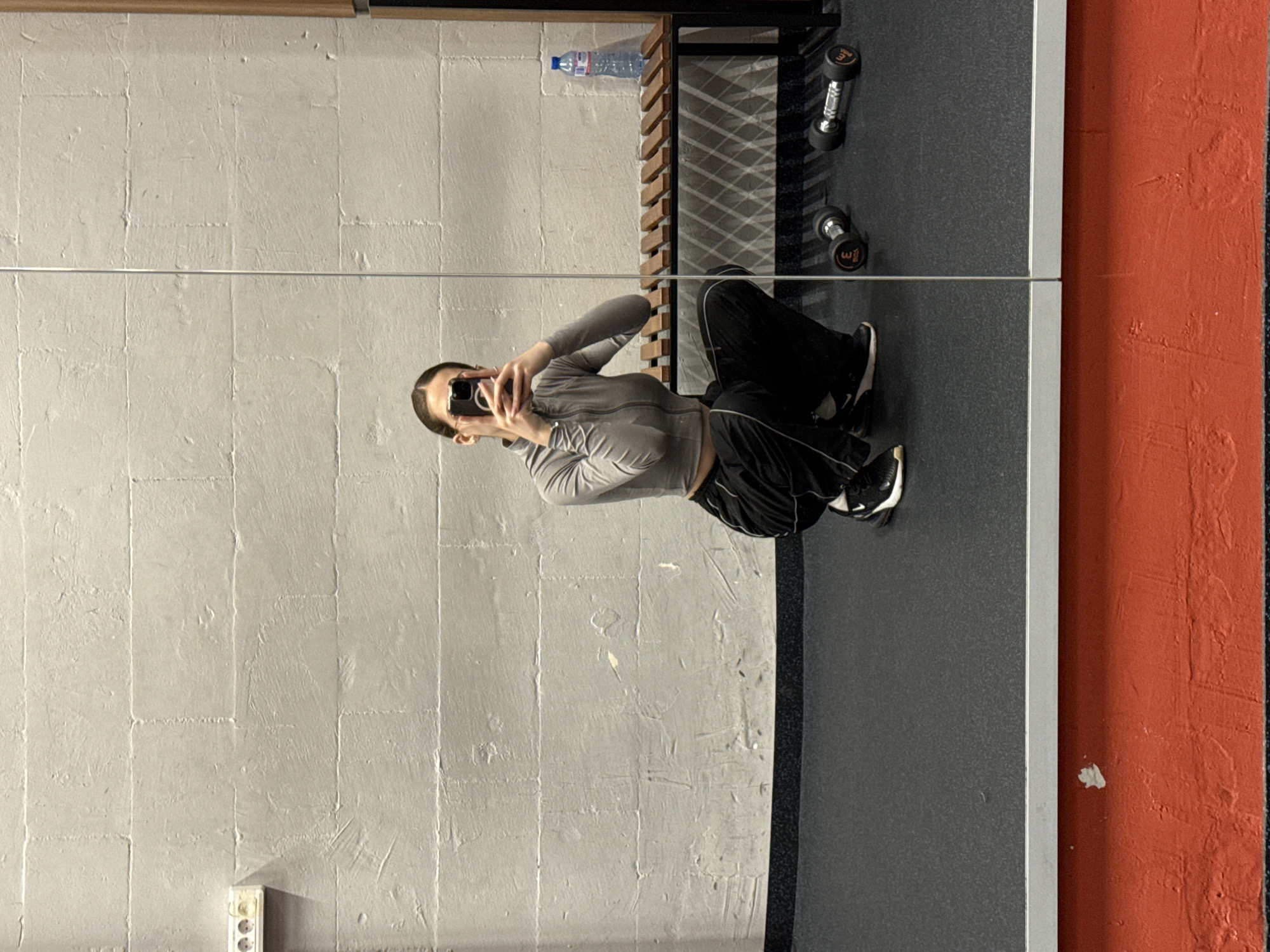 A woman is taking a selfie in a gym. She is wearing a gray zip-up jacket and black workout pants. The gym has gray brick walls and a gray floor with a red accent wall. Dumbbells and a water bottle are visible on a shelf in the background. A power outlet is seen on the wall. The location is possibly in Sofia, Bulgaria, but this cannot be confirmed definitively from the image.