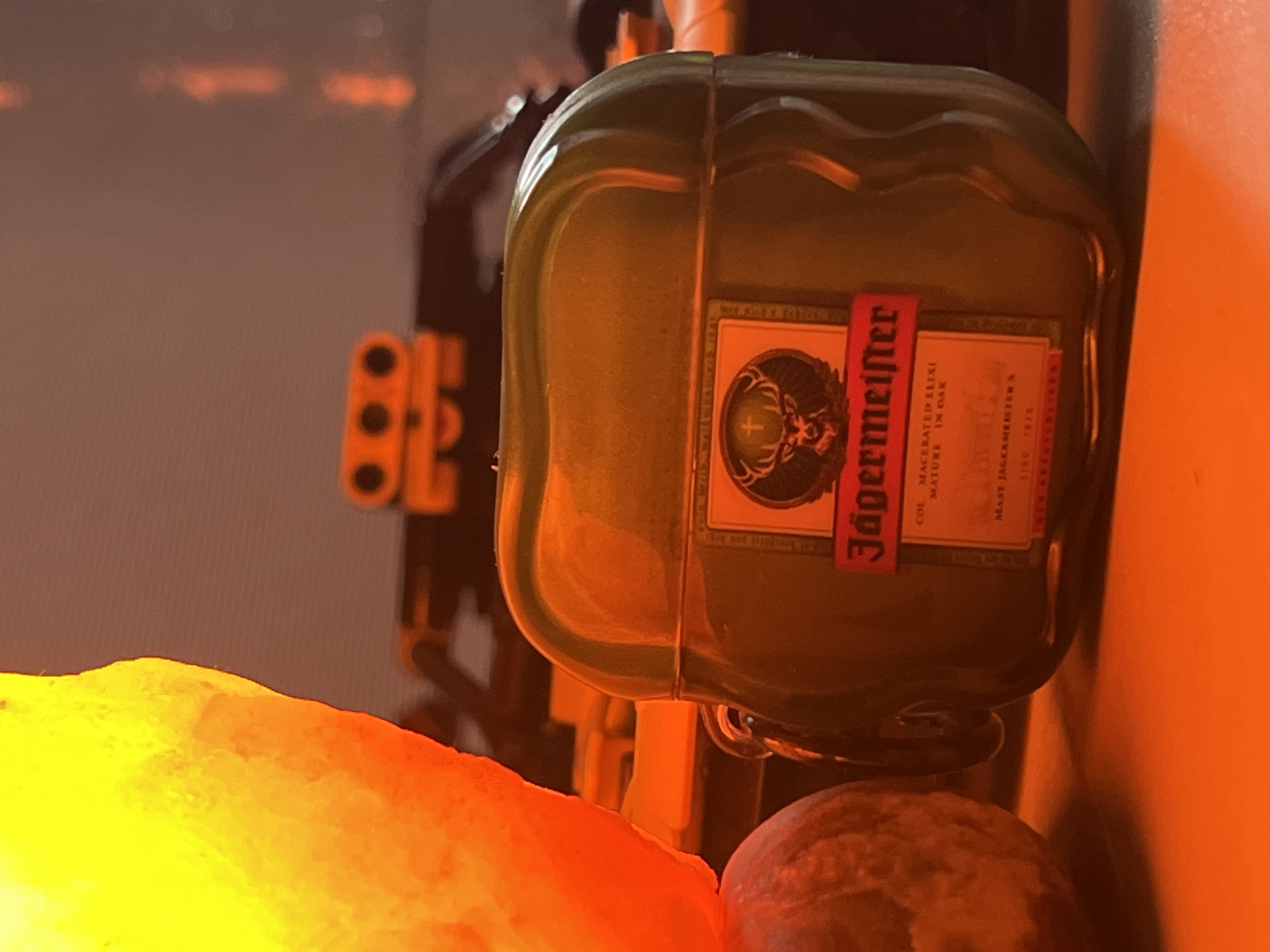 The image captures a close-up, still-life arrangement dominated by a dark green protective case, likely for earphones, styled to resemble a miniature Jägermeister liquor bottle. The case features a distinct wavy top edge and a red and white label. Visible text on the label includes "Jägermeister", "COL. MACERATED ELIXI", "MATURE IN OAK", "MAST JÄGERMEISTER S", and "SINC 1878", along with the brand's iconic stag and cross logo.
The scene is bathed in warm, ambient light, creating a cozy and dim atmosphere, suggesting evening or nighttime. To the left of the Jägermeister case, a large, irregularly shaped object glows brightly with intense yellow and orange light, indicative of a salt lamp. A round, textured object with a reddish-brown hue is partially visible in the lower right foreground. In the blurry background, behind the main subject, dark shapes and discernible orange and black elements suggest the presence of building blocks, possibly from a Lego construction.
No specific activity or event is taking place; the image presents a static display of personal and decorative items on what appears to be a desk or shelf. The warm lighting contributes to a relaxed setting.
Location context: Unknown, Unknown