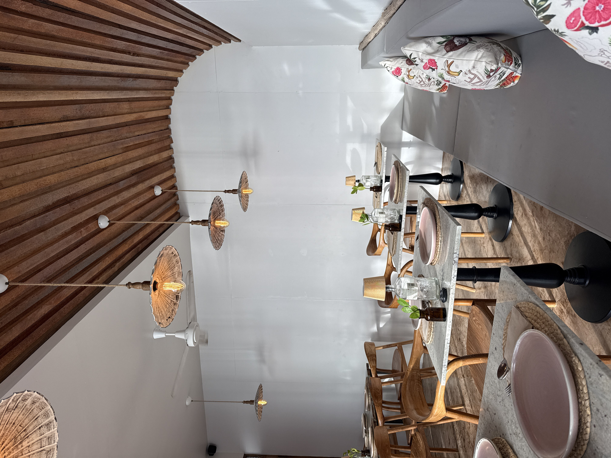 The image depicts the interior of a restaurant in Ko Samui District, Thailand. The restaurant features a modern, minimalist design with a focus on natural materials. The ceiling is constructed of wooden slats that curve inwards, and several pendant lights with woven shades hang from the ceiling. White walls provide a clean backdrop.
The restaurant is furnished with light-colored granite-top tables and wooden chairs with curved backs. Some tables are set with plates, cutlery, and small decorative vases. In the foreground, a close-up of a table shows a pink plate, a fork, and a spoon on a woven placemat.
A long, grey upholstered bench with patterned throw pillows is visible along the right side of the room. The pillows feature a tropical-themed print with fruits and flowers. A white ceiling fan is mounted on the wall above the tables. Natural light streams in, creating shadows on the walls. The overall impression is one of a stylish and serene dining environment. No people are visible in the image.