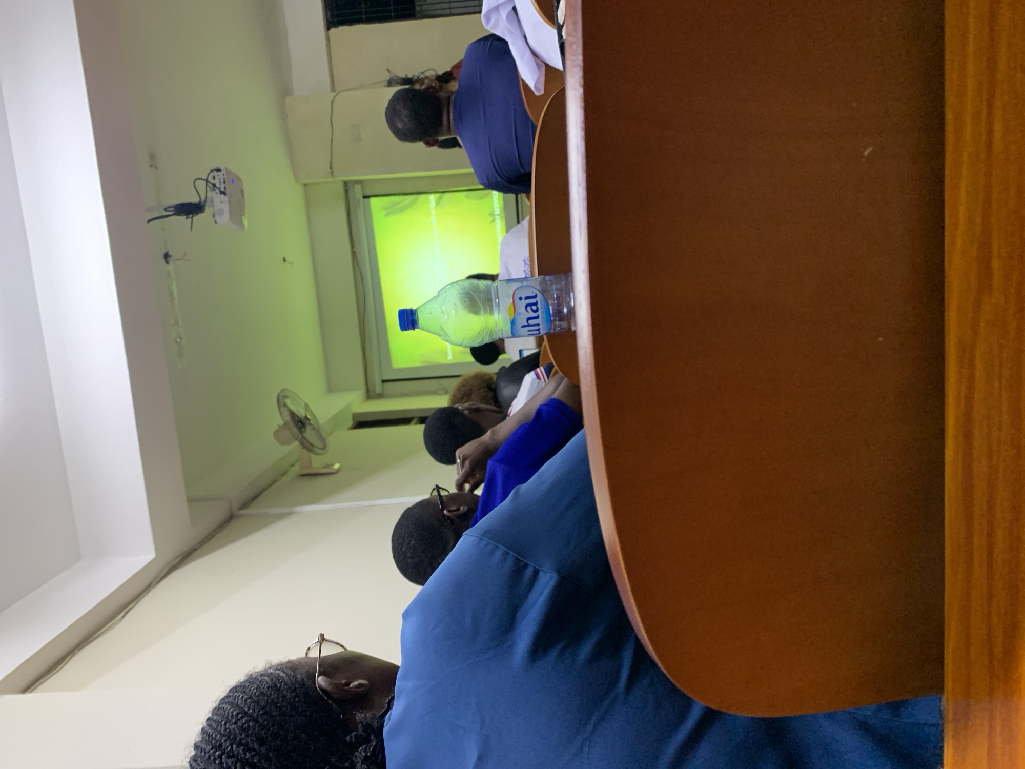 A classroom in Dar es Salaam, Tanzania, is shown with students seated at desks facing a projection screen. A water bottle labeled "Uhail" sits on a desk. A ceiling fan is visible. A projector is mounted on the wall. Most students appear to be focusing on the presentation.