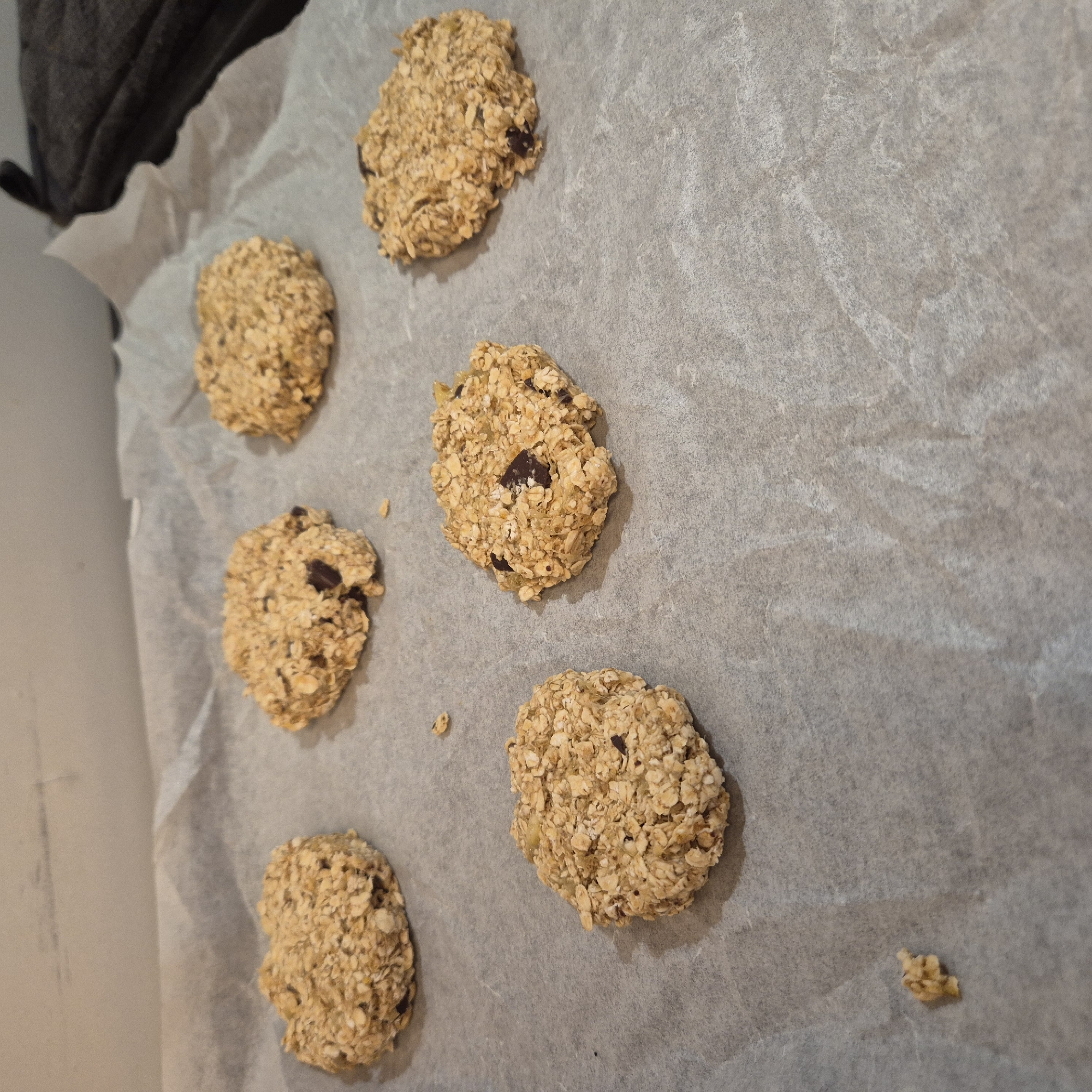 The image shows a baking sheet lined with parchment paper on which there are six irregularly shaped oatmeal cookies, some with visible chocolate chips. A small clump of oatmeal is near the bottom right of the sheet. The location and city of Strasbourg, France, cannot be determined from the image. No people, interactions, or specific environment can be identified.