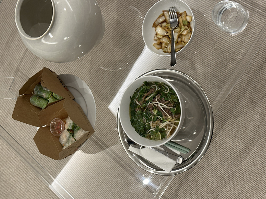 A table is set with a grey ceramic vase, two open cardboard boxes containing spring rolls with dipping sauce, a bowl of stir-fried vegetables with a fork, a glass of water, and a bowl of pho on a silver tray with chopsticks and cutlery. The scene is brightly lit, suggesting daytime. The table has a clear, reflective surface, possibly glass.