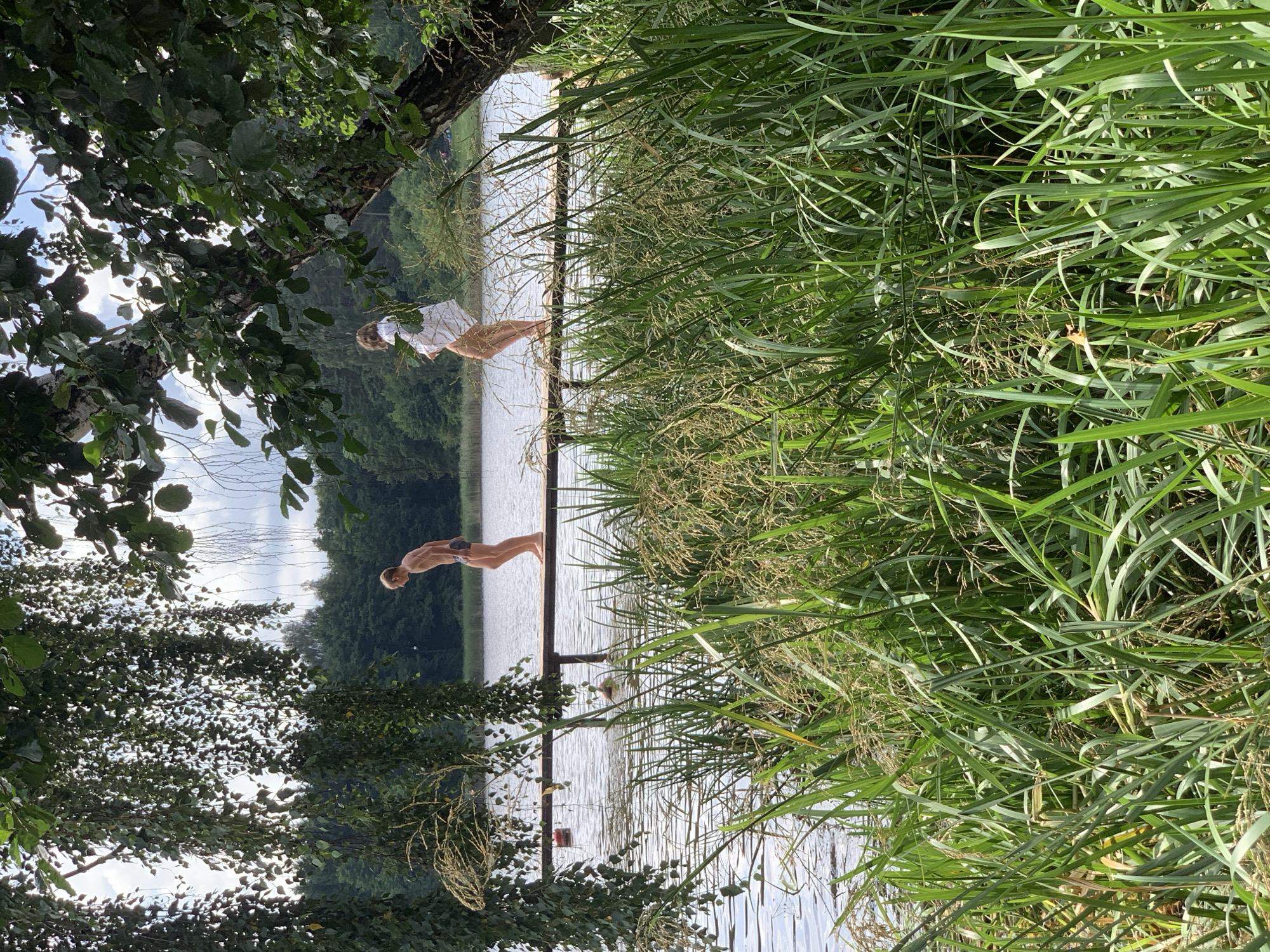 Two individuals are present on a wooden pier extending over a body of water. The person on the left, a male, is standing with his back to the camera, facing the water. He is wearing dark-colored swim trunks. The person on the right, a female, is also standing and facing away from the camera, appearing to be looking out at the water or the horizon. She is wearing a white garment, possibly a shirt or dress, and her legs are visible. In the foreground, lush green reeds and tall grass grow densely along the water's edge, obscuring some of the lower portion of the pier and the water. The background features a dense line of trees, suggesting a natural, possibly forested, environment. The sky above is partly cloudy. The scene appears to be set in a natural outdoor location, likely a lake or a wide river, within the Грозаўскі сельскі Савет in Belarus, based on the provided location context.