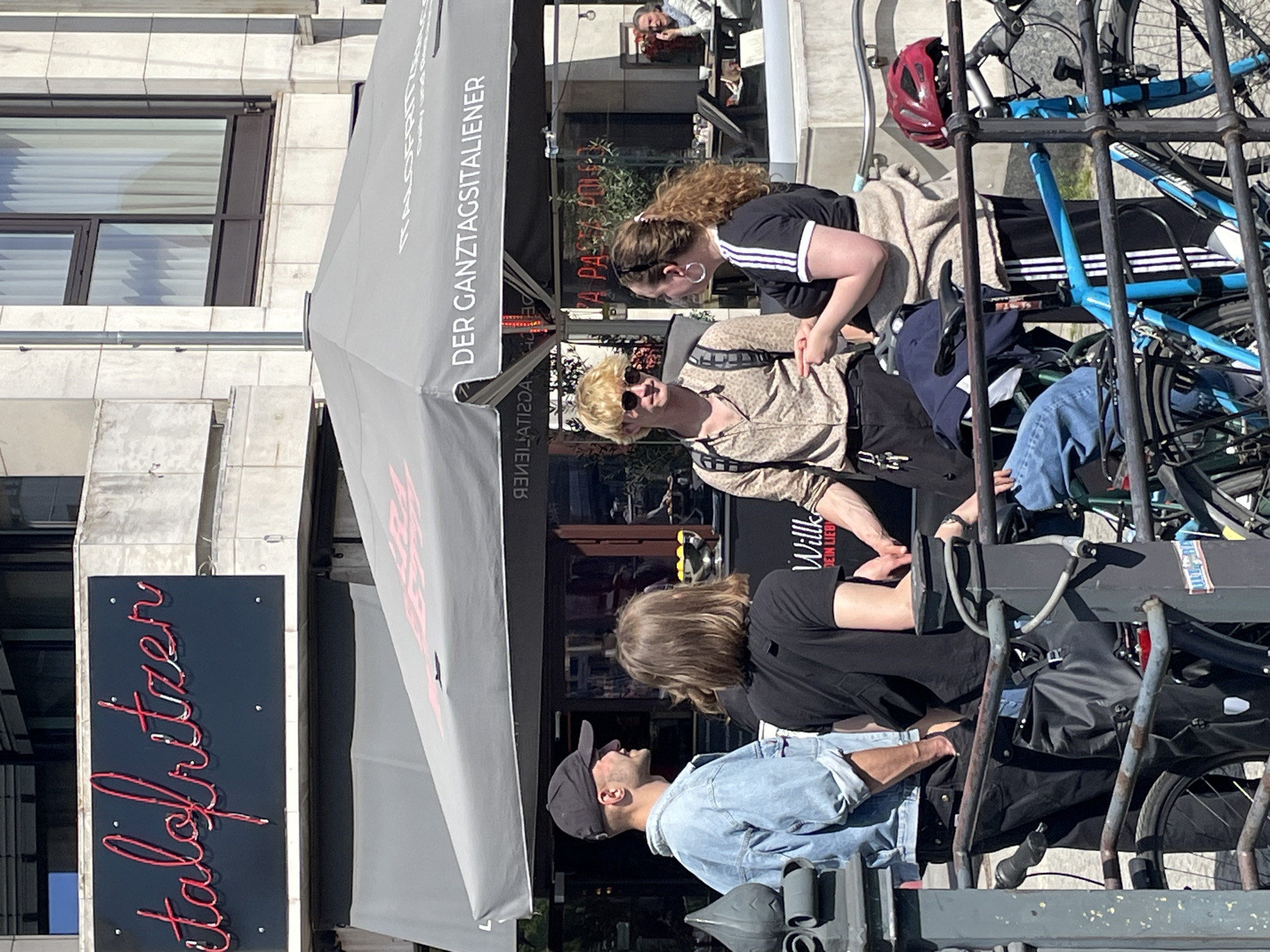 This outdoor urban scene, set in Berlin, Germany, depicts several people gathered around an establishment, possibly a cafe or restaurant, under a large grey canopy. The time of day appears to be bright daylight, suggesting fair weather.
In the foreground, on the right side, a sturdy metal bicycle rack holds multiple bicycles, including a prominent blue one with a red helmet resting on its handlebars. A person wearing a black baseball cap and a denim jacket is visible in the bottom right, looking upwards and to the right.
Toward the center, under the grey canopy, are several individuals. A person with blonde hair, wearing sunglasses and a patterned shirt, is looking towards the right. Beside them, another person with dark curly hair and a black t-shirt with white stripes is looking downwards. A third person with brown hair and a black t-shirt is partially visible behind them. The people appear to be socializing or relaxing.
The grey canopy bears German text, "DER GANZTAGSITALIENER," which translates to "The All-Day Italian," indicating the type of establishment. Above this, "KA...FRITZ..." is partially visible, likely part of the name. Behind the canopy, through large glass windows, parts of the building interior are visible, including what looks like a counter or bar area and another person inside. "A PASTA" is also visible through the window.
On the far left, a vertical black sign with red neon script displays "talfritzen," suggesting another business nearby, perhaps "Gaststätte Fritz." The building facade on the left is made of light-colored stone or concrete with windows featuring horizontal blinds.
Overall, the image captures a casual street scene with people enjoying the outdoors, surrounded by typical urban elements like buildings, bicycles, and commercial signage.