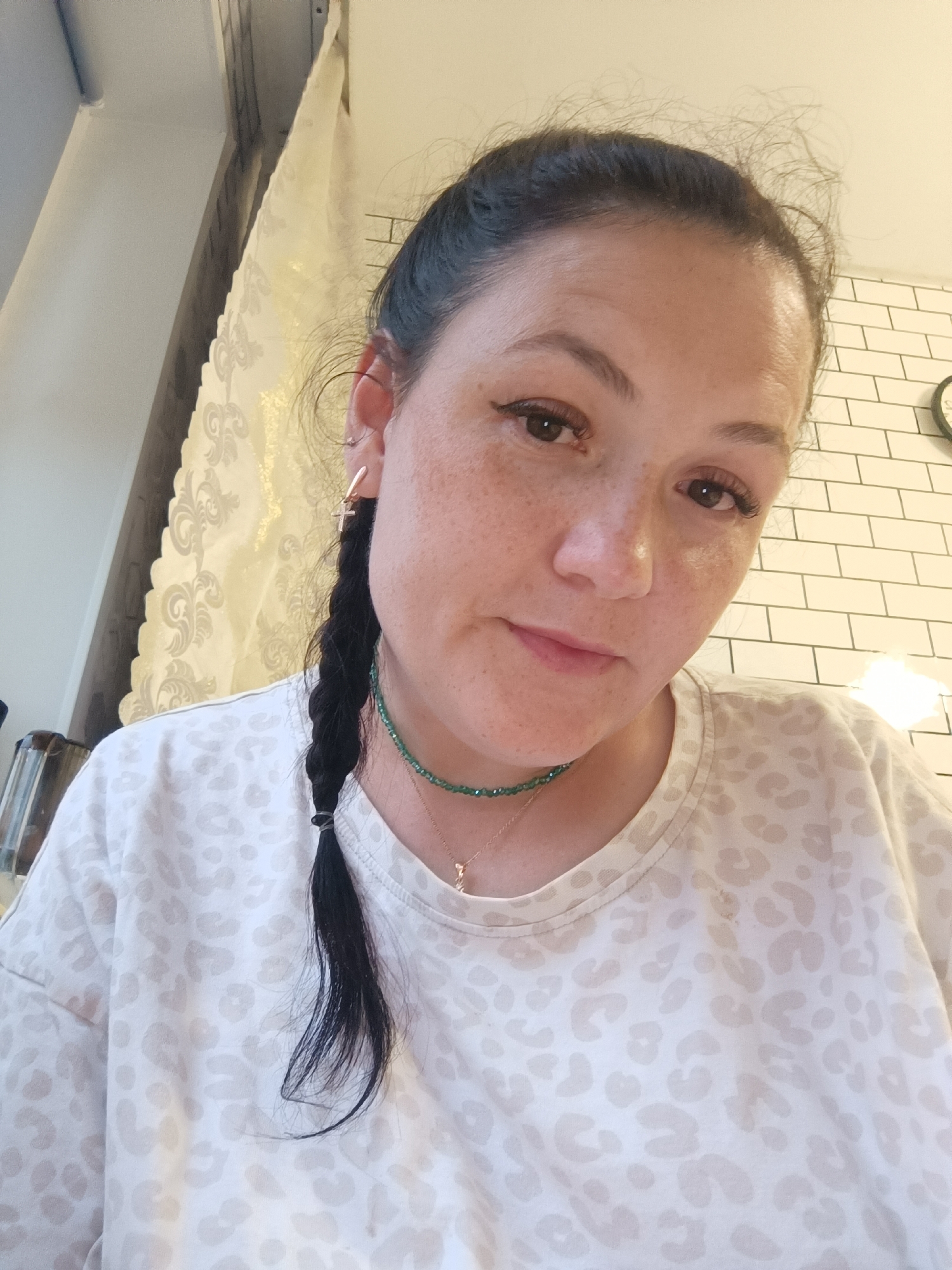 A woman with dark hair in a braid, wearing a light-colored, leopard-print shirt and a green beaded necklace, looks directly at the camera. She has light skin with freckles and appears to be wearing subtle makeup. The background includes a white tiled wall and what looks like a window with a decorative curtain. The setting is indoors, with artificial lighting. No specific location in Chaikovsky, Russia, is identifiable from the image.