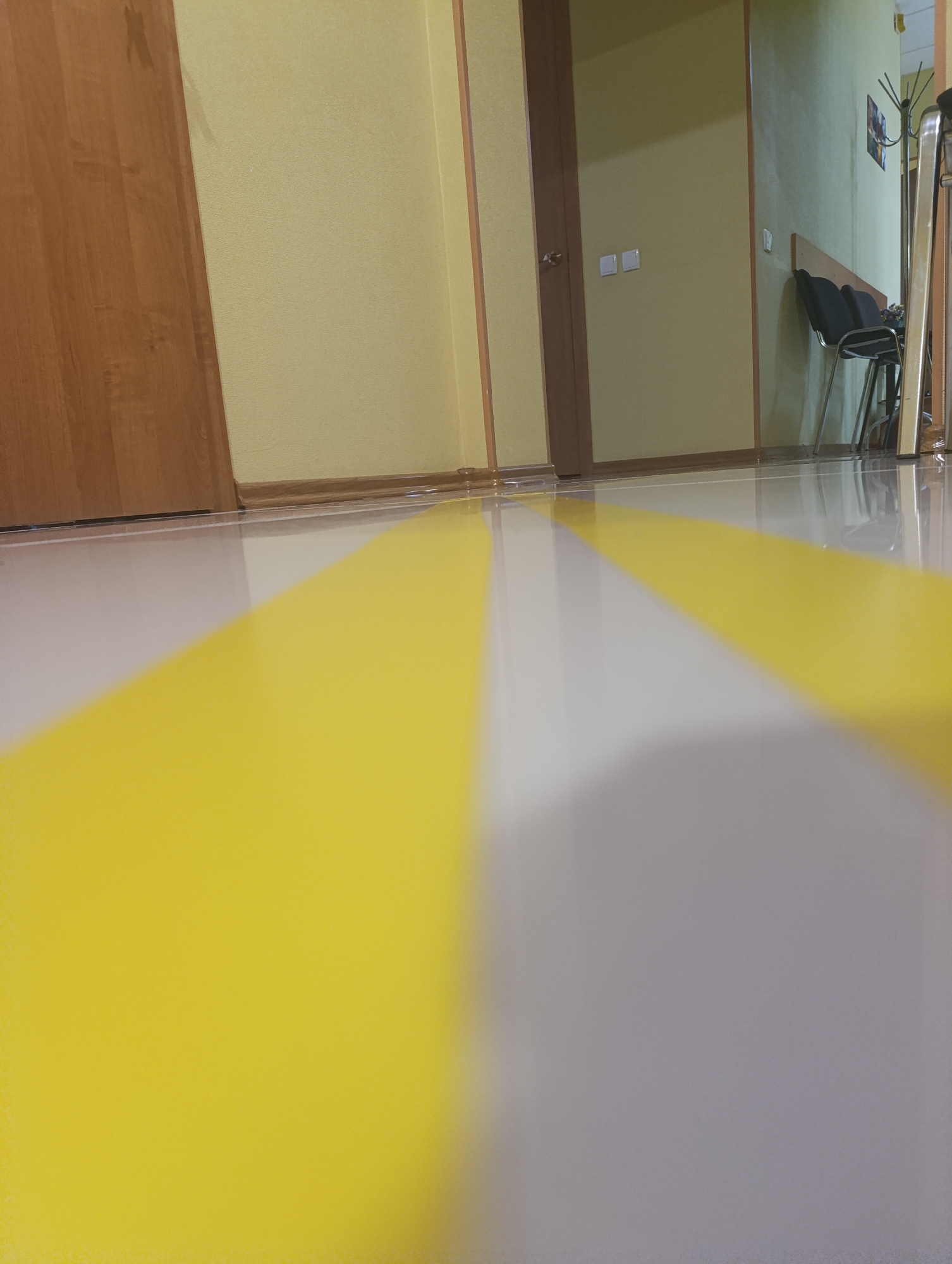 In an indoor hallway, a low-angle, wide shot captures a reflective, glossy floor with diagonal yellow and white stripes. To the left, a tall, light-brown wooden door stands closed. The walls are painted a pale yellow. In the background, a dark brown door with a doorknob is visible, next to a pale yellow wall with two white light switches. Further down the hallway, two black chairs are positioned in front of a pale green wall. A coat rack with items on it and a railing of a staircase are also present in the background. The scene appears to be in an office or public building, possibly a waiting area.