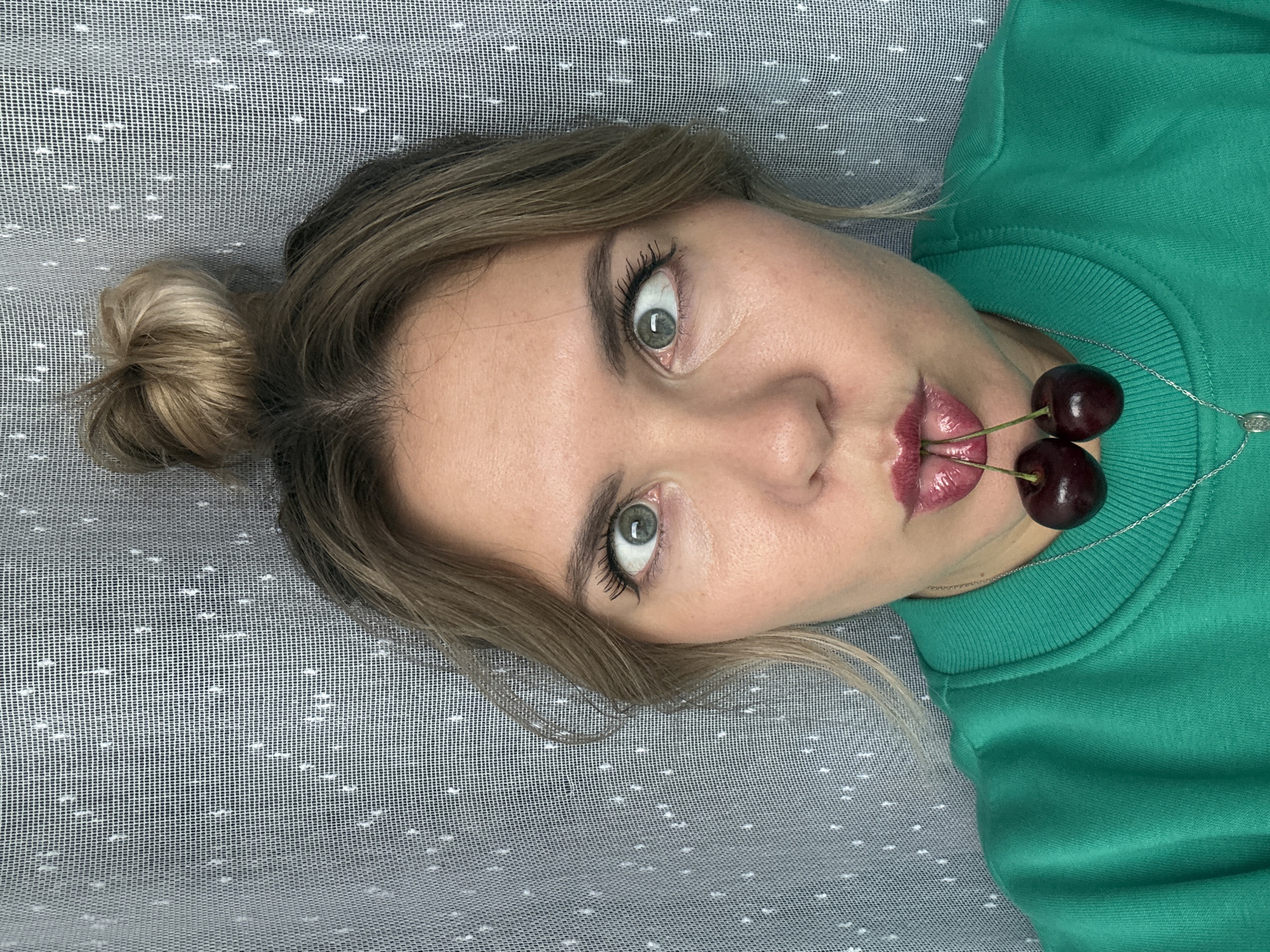 A young woman with light brown hair pulled into a bun at the top of her head, is looking directly at the camera with a playful expression. She is wearing a bright green sweatshirt and has two cherries held between her lips. Her eyes are wide and crossed, adding to the humorous effect. The background is a grey curtain with a subtle white dot pattern. The image appears to be a selfie taken indoors, likely in a residential setting. There is no specific information in the image to suggest a location in Izhevsk, Russia.