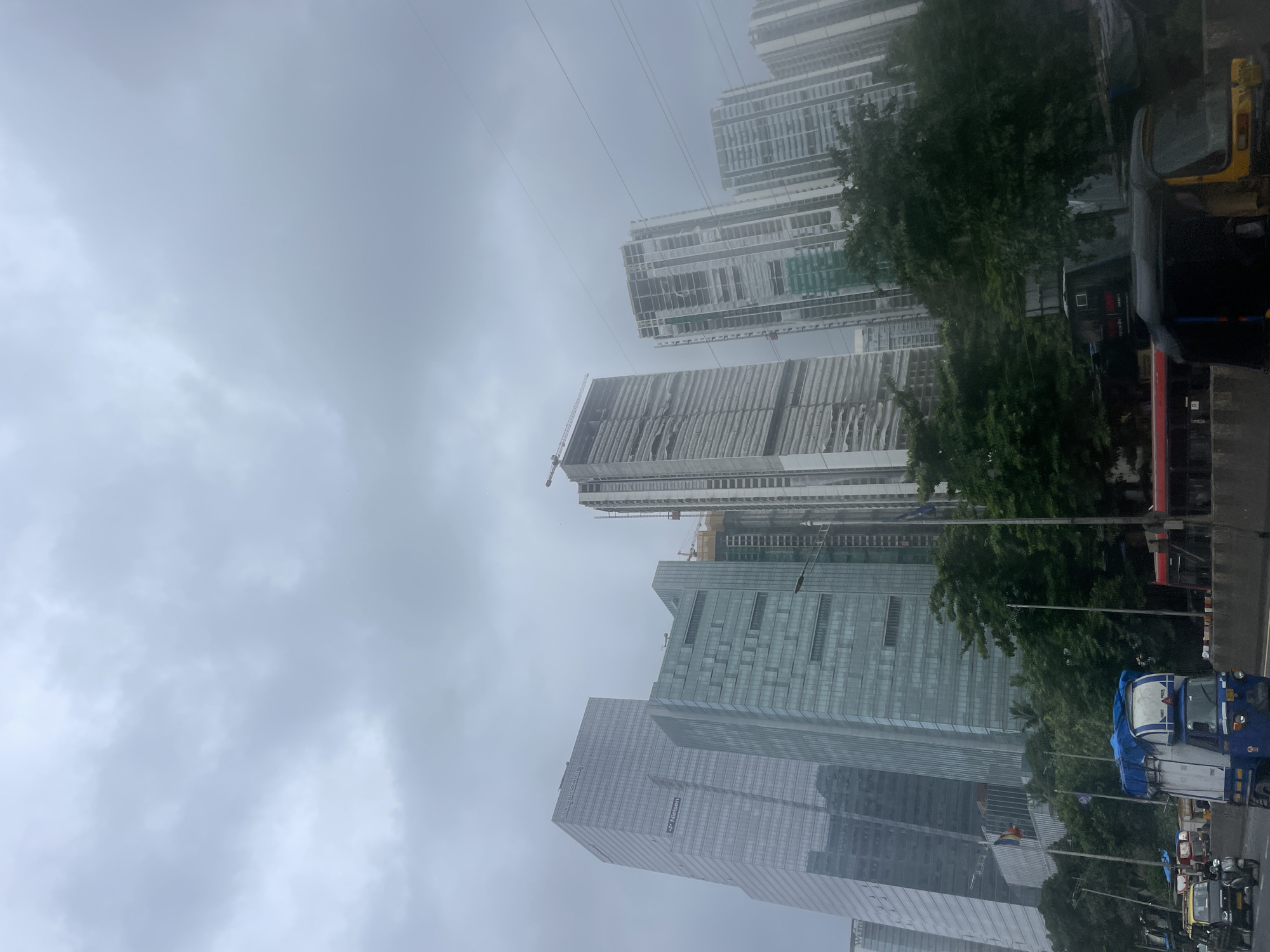 In Mumbai, India, on a cloudy day, a perspective shot from a street level shows a cluster of modern skyscrapers, some under construction, dominating the cityscape. The buildings feature glass facades, with visible signage for "Morgan Stanley" and "Nielsen" on one of the completed towers. The foreground is occupied by a road with several vehicles, including an auto-rickshaw, a yellow taxi, and a truck with a blue tarp covering its cargo. Lush green trees line the median of the road, separating the traffic lanes. In the background, construction cranes are visible against the overcast sky. The scene captures a typical urban environment in Mumbai, characterized by a mix of contemporary architecture and local transportation.
