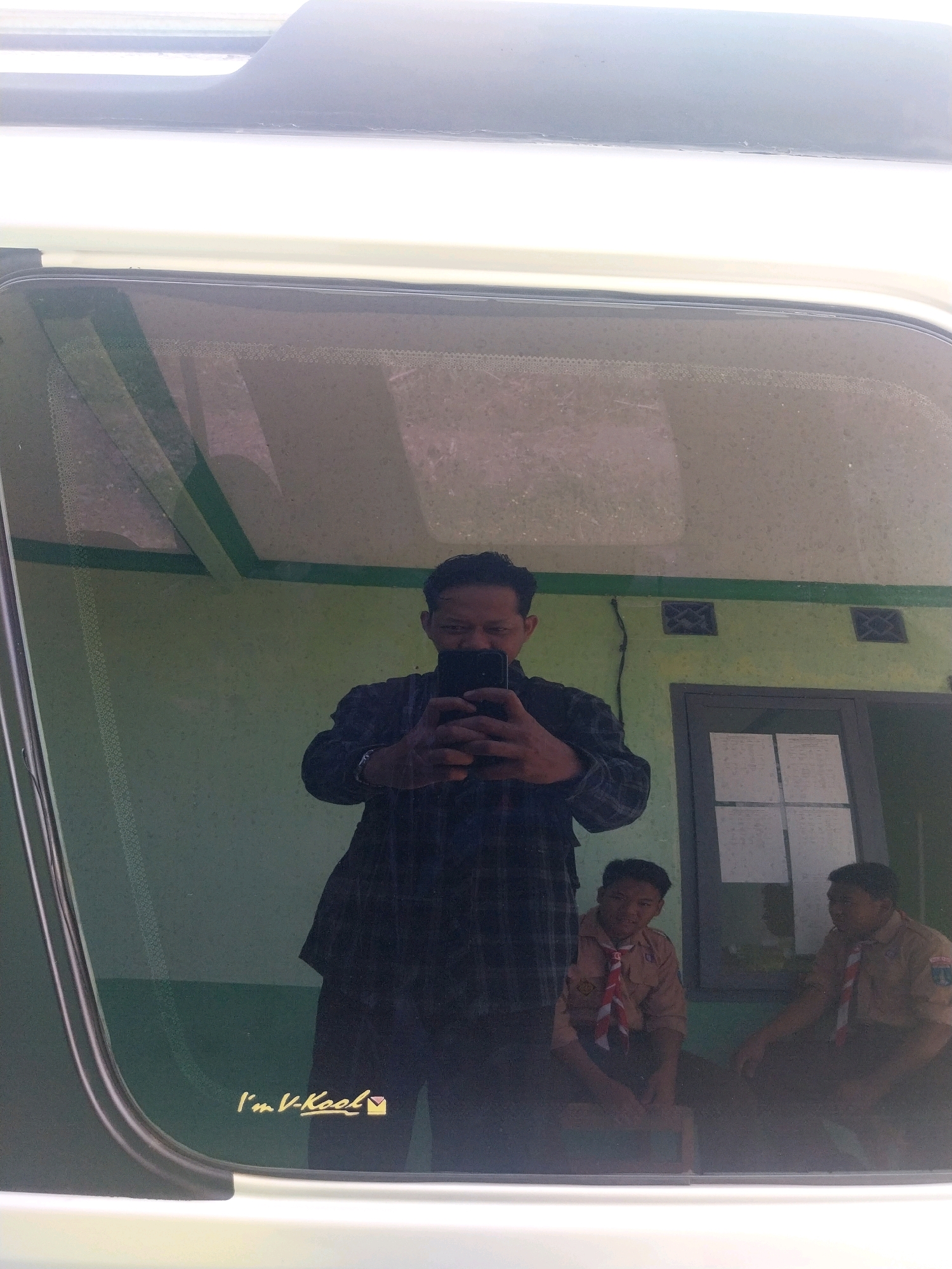 The image captures a reflection in a vehicle window, offering a glimpse of a street scene in Pandean, Indonesia. In the foreground, a man in a plaid shirt is reflected as he takes a selfie with his phone. Behind him, two boys in scout uniforms are also visible in the reflection. The background shows a light green wall with two square vents and a window with papers inside. A sticker with the text "I'm V-Kool" is present on the lower left side of the vehicle window. The scene suggests an outdoor setting on a sunny day, likely in a residential or school area.