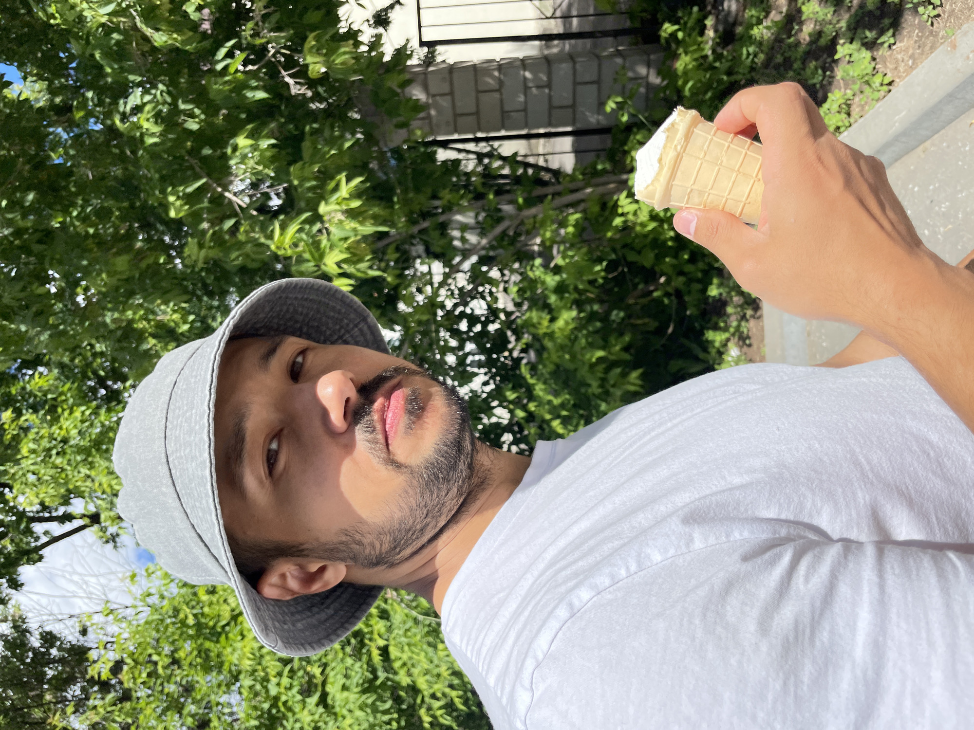A man in a white t-shirt and grey bucket hat holds an ice cream cone. The background shows green foliage and a building. The location is not discernible as Izhevsk.